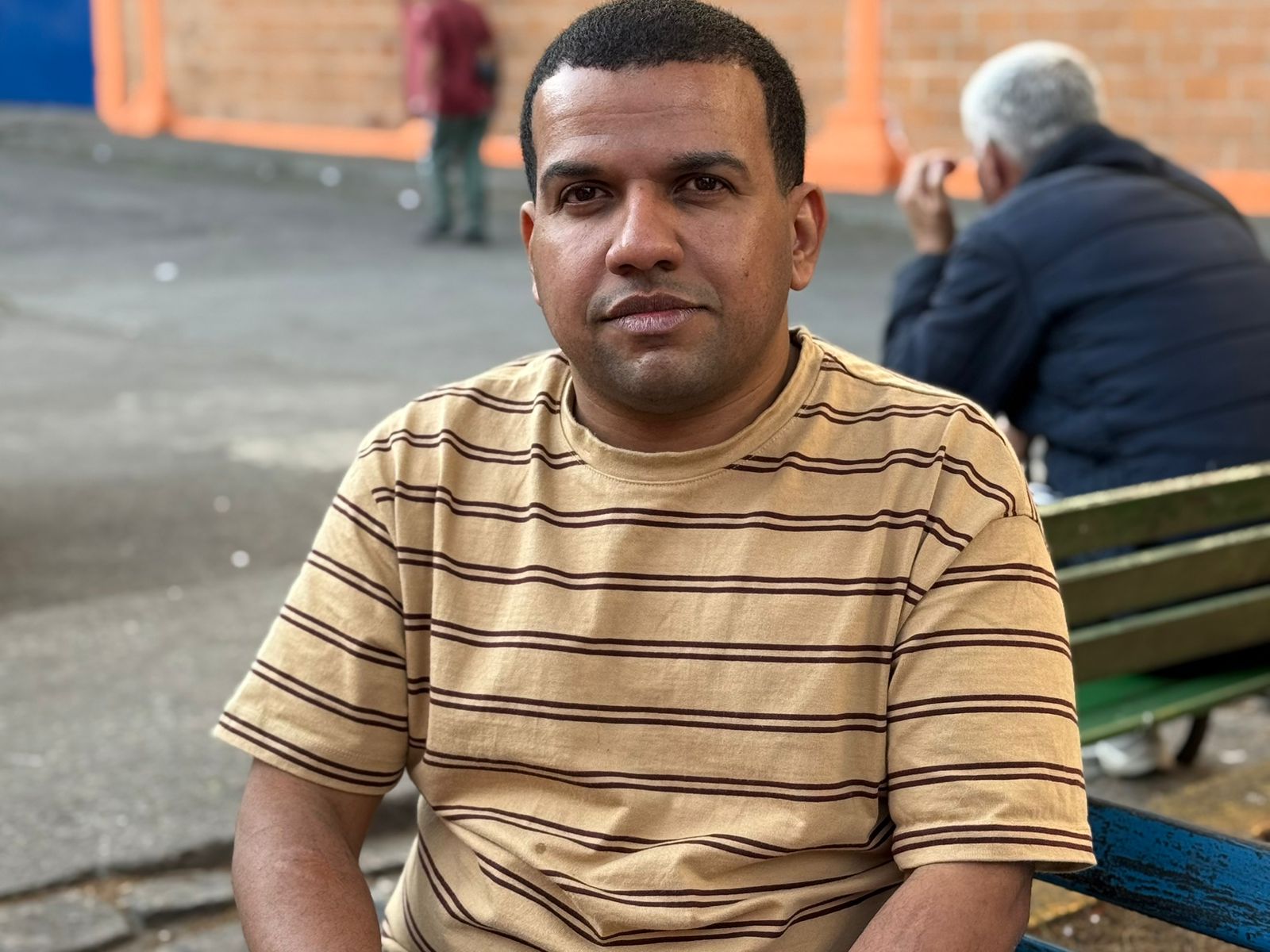 Lucas Amaral sentado em banco ao ar livre, olhando para a câmera, em ambiente urbano com outras pessoas ao fundo.