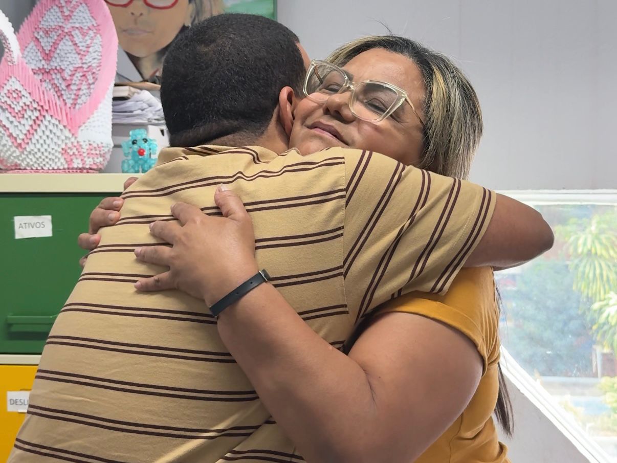 Lucas e Ivone se abraçam em ambiente interno, transmitindo acolhimento e afeto, com armários coloridos ao fundo.
