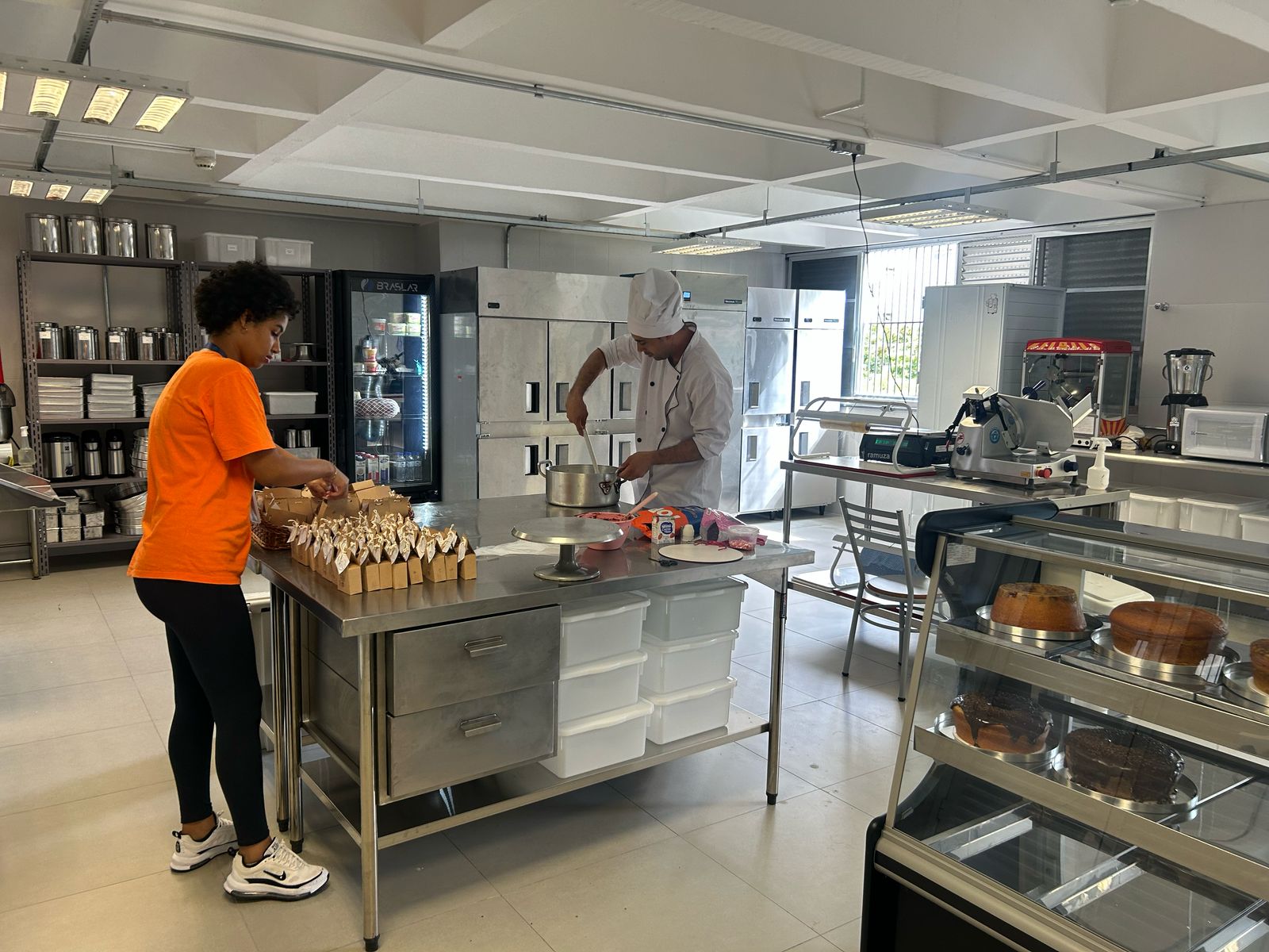 A imagem mostra uma cozinha industrial bem equipada, com bancadas de aço inoxidável, armários, refrigeradores e prateleiras com potes organizados. No ambiente, duas pessoas estão em atividade.  Uma mulher vestindo uma camiseta laranja e calça preta está organizando pequenas embalagens sobre a mesa, que parecem conter alimentos prontos para distribuição. Ela está de tênis brancos e parece concentrada na tarefa.  Ao fundo, um homem vestindo uniforme de chef, incluindo um avental branco e um chapéu de cozinheiro, está mexendo o conteúdo de uma panela sobre uma bancada.