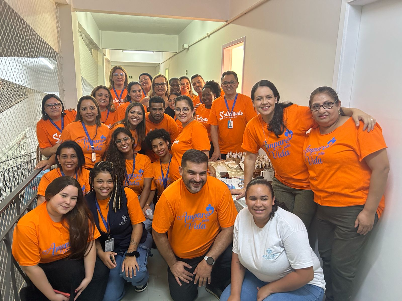 A imagem mostra um grupo de aproximadamente 20 pessoas reunidas em um corredor, posando para uma foto. A maioria está vestindo camisetas laranjas com inscrições, e algumas pessoas usam crachás. Há também uma mulher vestindo branco. Todos parecem felizes, com expressões sorridentes, e algumas pessoas estão abraçadas ou apoiando as mãos nos ombros umas das outras.