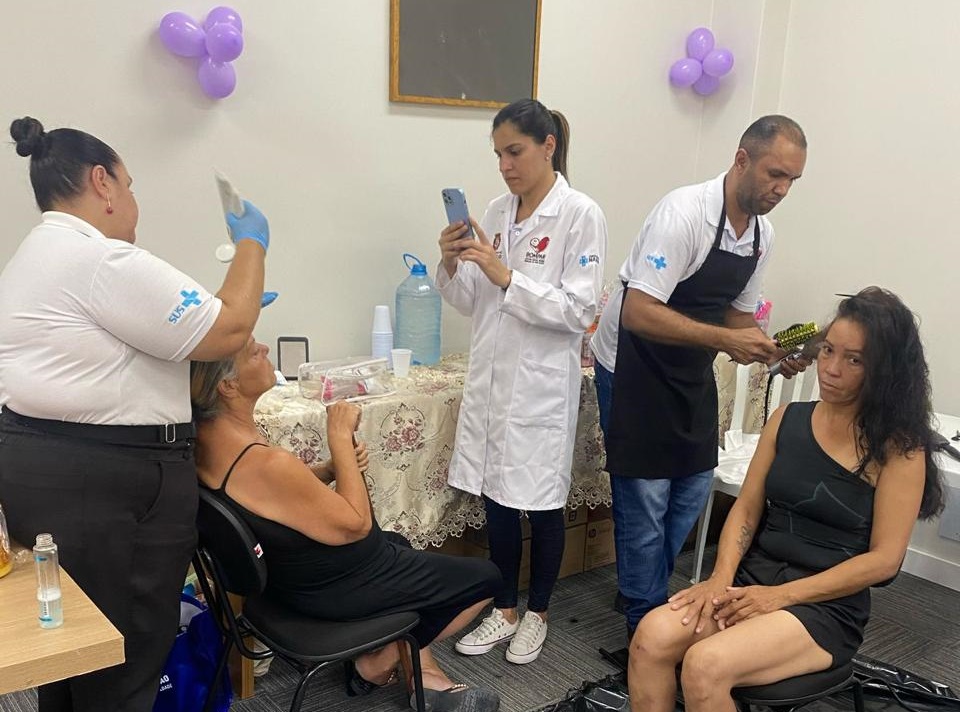 Em uma sala com decoração simples, balões roxos e uma mesa coberta com toalha rendada, profissionais realizam atendimentos de beleza e saúde. À esquerda, uma funcionária do SUS, de uniforme branco, usa luvas e aplica um produto no rosto de uma mulher sentada. Ao centro, uma profissional de jaleco branco registra algo no celular. À direita, um cabeleireiro de avental preto escova os cabelos de uma mulher vestida de preto, que está sentada em um banco.