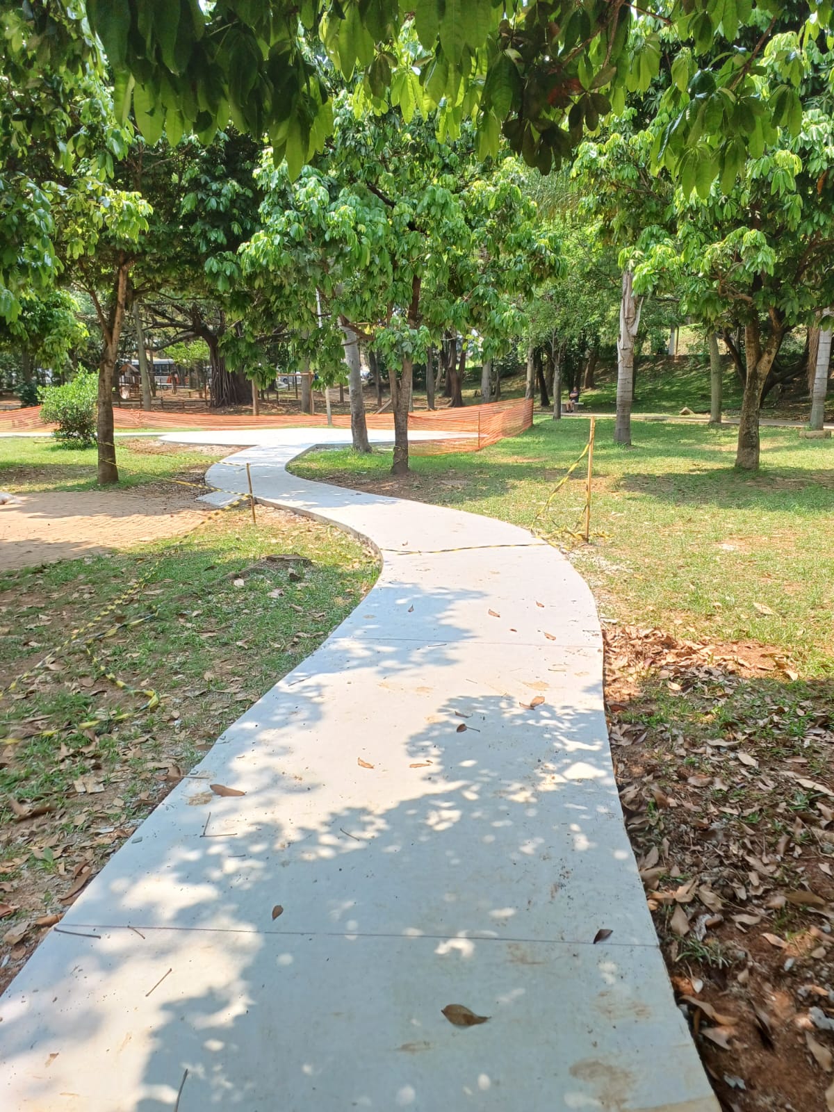 Foto diurna em plano médio, que captura um caminho cimentado novo, de cor clara e superfície lisa, que se curva suavemente, estendendo-se do primeiro plano para o fundo da imagem. O caminho está ladeado por grama verde, terra avermelhada e folhas secas. A praça é densamente arborizada, com muitas árvores de troncos finos e copas cheias, que projetam sombras no piso. No fundo, é possível ver uma cerca de segurança laranja (indicando que a área pode estar em reforma) e mais vegetação. O céu é parcialmente visível, sugerindo um dia ensolarado.