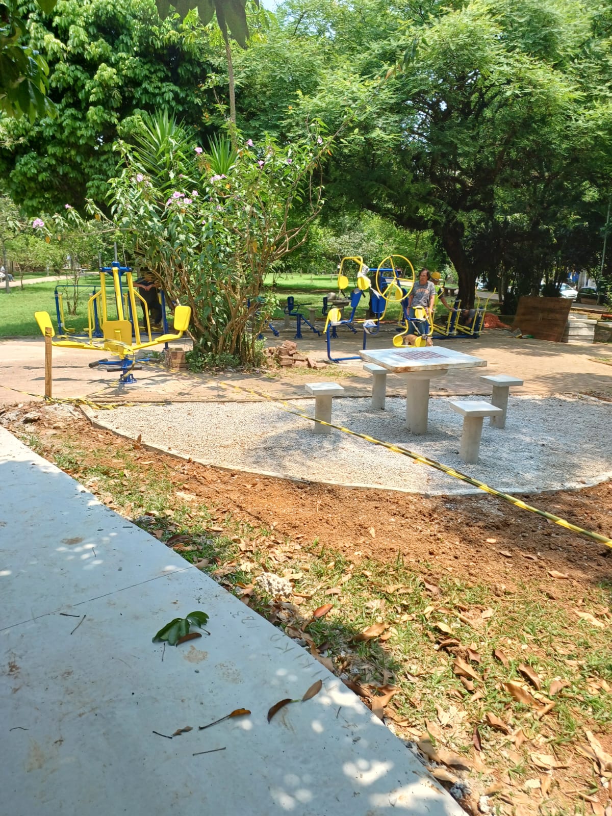 Foto diurna em plano médio de uma área recém-revitalizada na praça. Em primeiro plano, vê-se o final de um caminho cimentado e, à frente, uma área circular com piso de pedrisco claro, delimitada por fita de advertência (amarela e preta). No centro dessa área, há um conjunto de mesas e bancos de concreto. Logo atrás, em um piso de terra batida, estão os aparelhos de ginástica (Academia ao Ar Livre) pintados de amarelo e azul, cercados por folhagens. Uma pessoa está sentada em um dos aparelhos no fundo. O sol brilha intensamente, e a área é cercada por árvores grandes e muita vegetação.