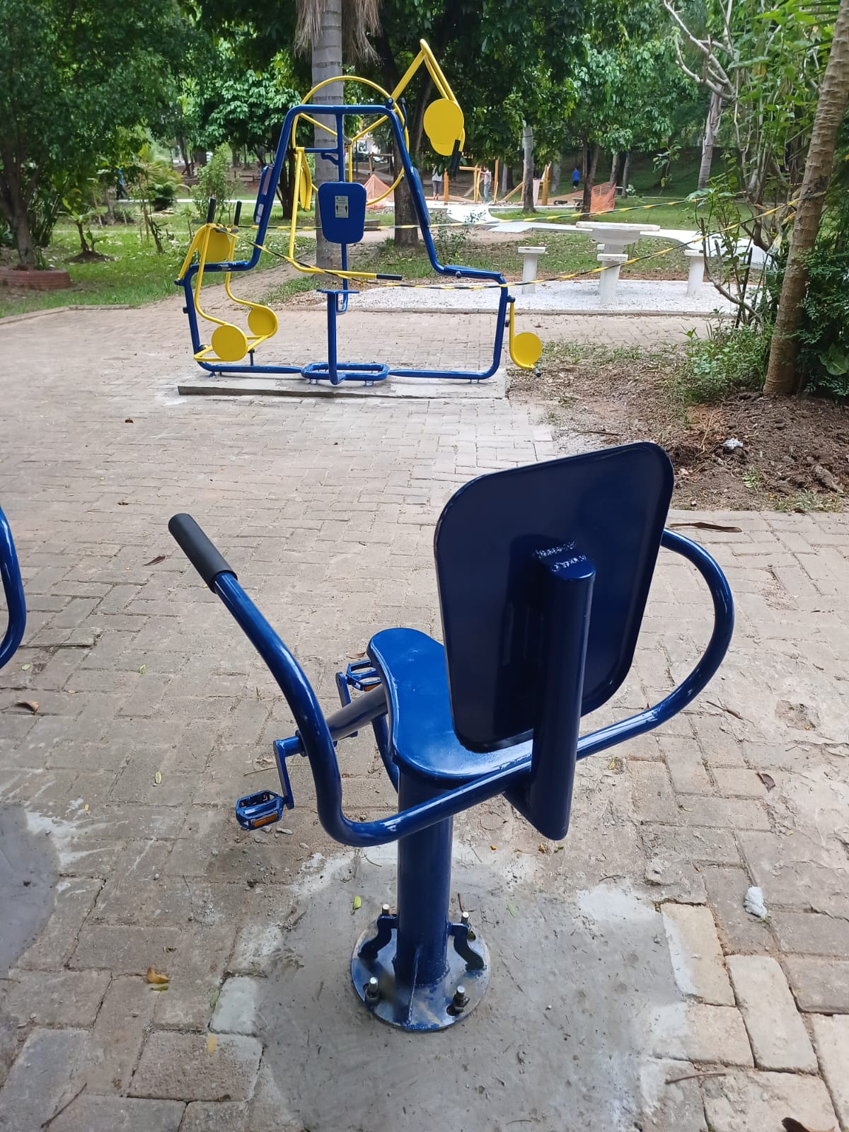 Foto diurna em close de dois aparelhos da Academia ao Ar Livre. Em primeiro plano, um aparelho de bicicleta reclinada de cor azul escura e brilhante, com um assento e encosto bem definidos. O equipamento está fixado em um piso de blocos de concreto intertravados. No segundo plano, um outro aparelho de ginástica (provavelmente para pernas/caminhada) em azul e amarelo se destaca. Ao fundo, parcialmente visível, está a nova área de mesas e bancos de concreto. O ambiente é sombreado por árvores.