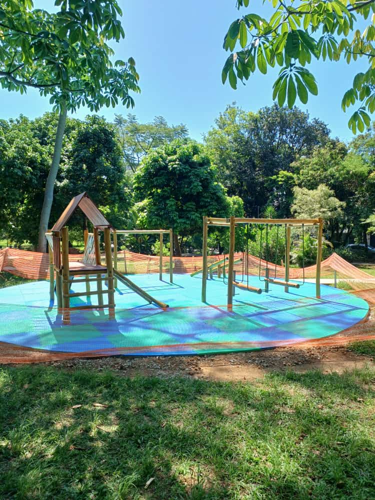Foto diurna de um parquinho infantil recém-reformado, localizado em uma área gramada e arborizada. O parquinho possui uma estrutura moderna de madeira clara, com um pequeno forte, escorrega e balanços. O destaque é o piso emborrachado multicolorido, em tons de azul, verde e roxo, formando padrões geométricos. Uma cerca de segurança temporária (laranja) envolve parcialmente a área do parquinho. O dia está ensolarado e o céu é azul.