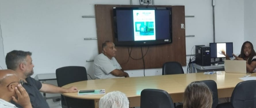 A foto mostra um grupo de pessoas reunidas em uma sala, participando de uma reunião ou apresentação. A sala é iluminada por luz artificial e tem paredes claras.  No lado esquerdo da foto, está o Subprefeito da Casa Verde/Limão/Cachoeirinha Eduardo Valentim,um homem de pele morena clara sentado em uma cadeira, de costas para a câmera, com a mão esquerda na mesa. Ele parece estar ouvindo atentamente. Ao seu lado, um homem de pele clara, vestindo uma camisa branca, está sentado à mesa, de frente para a câmera. Ele também parece estar prestando atenção na apresentação.  No centro da foto, uma mulher de pele morena clara está sentada à mesa, de frente para a câmera. Ela está usando um laptop e parece estar falando ou apresentando algo. Ao seu lado, uma mulher de pele clara, com cabelos castanhos, está sentada à mesa, também de frente para a câmera. Ela está gesticulando com as mãos enquanto fala.  À direita, um homem de pele morena clara está sentado em uma cadeira, de costas para a câmera. Ele parece estar ouvindo a apresentação. Ao seu lado, um homem de pele clara, com cabelos grisalhos, está sentado à mesa, de frente para a câmera. Ele também parece estar prestando atenção.  Ao fundo, na parede, há uma televisão de tela plana ligada, exibindo um slide de apresentação. Ao lado da televisão, há um computador preto em cima de uma mesa menor.  No centro da sala, há uma mesa oval de madeira clara, onde as pessoas estão sentadas. As cadeiras são pretas, com encosto de tela.  Atrás das pessoas, há uma janela com persianas fechadas. A sala parece ser um ambiente de trabalho ou estudo, como uma sala de reuniões ou um auditório pequeno.