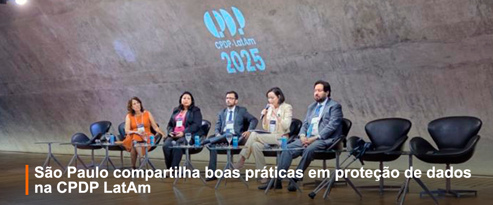 Foto registrando o evento, no palco 3 mulheres e dois homens, sentados, palestrando
