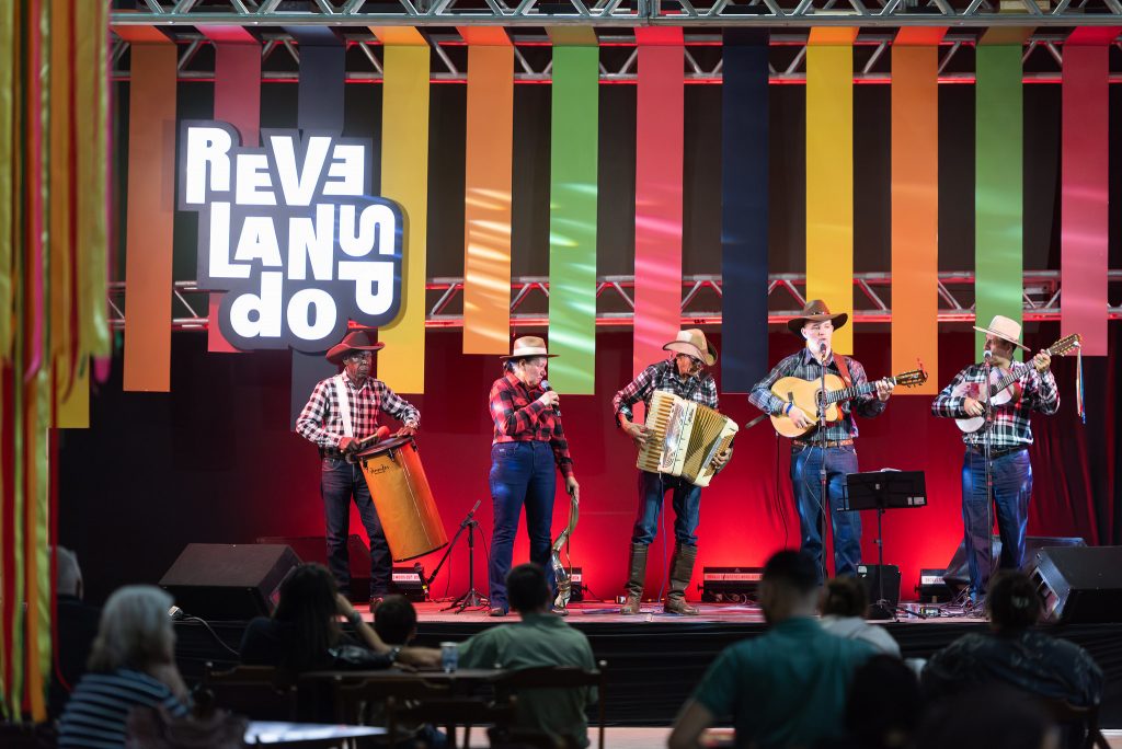 A imagem mostra um grupo musical se apresentando no palco durante o evento chamado "Revelando SP", conforme indicado pelo letreiro luminoso no fundo. A banda é composta por cinco músicos vestidos com trajes típicos caipiras, incluindo camisas xadrez, calças jeans e chapéus caipira.  Cada integrante toca um instrumento diferente: um segura uma caixa de folia (um tambor vertical), outro toca um acordeão, enquanto os demais tocam violão e viola caipira. O palco tem uma iluminação vibrante com luzes vermelhas e uma decoração de fitas coloridas penduradas na vertical, remetendo à cultura tradicional brasileira.  Na plateia, algumas pessoas estão sentadas assistindo à apresentação. O evento é voltado à valorização da cultura popular e tradicional do estado de São Paulo.