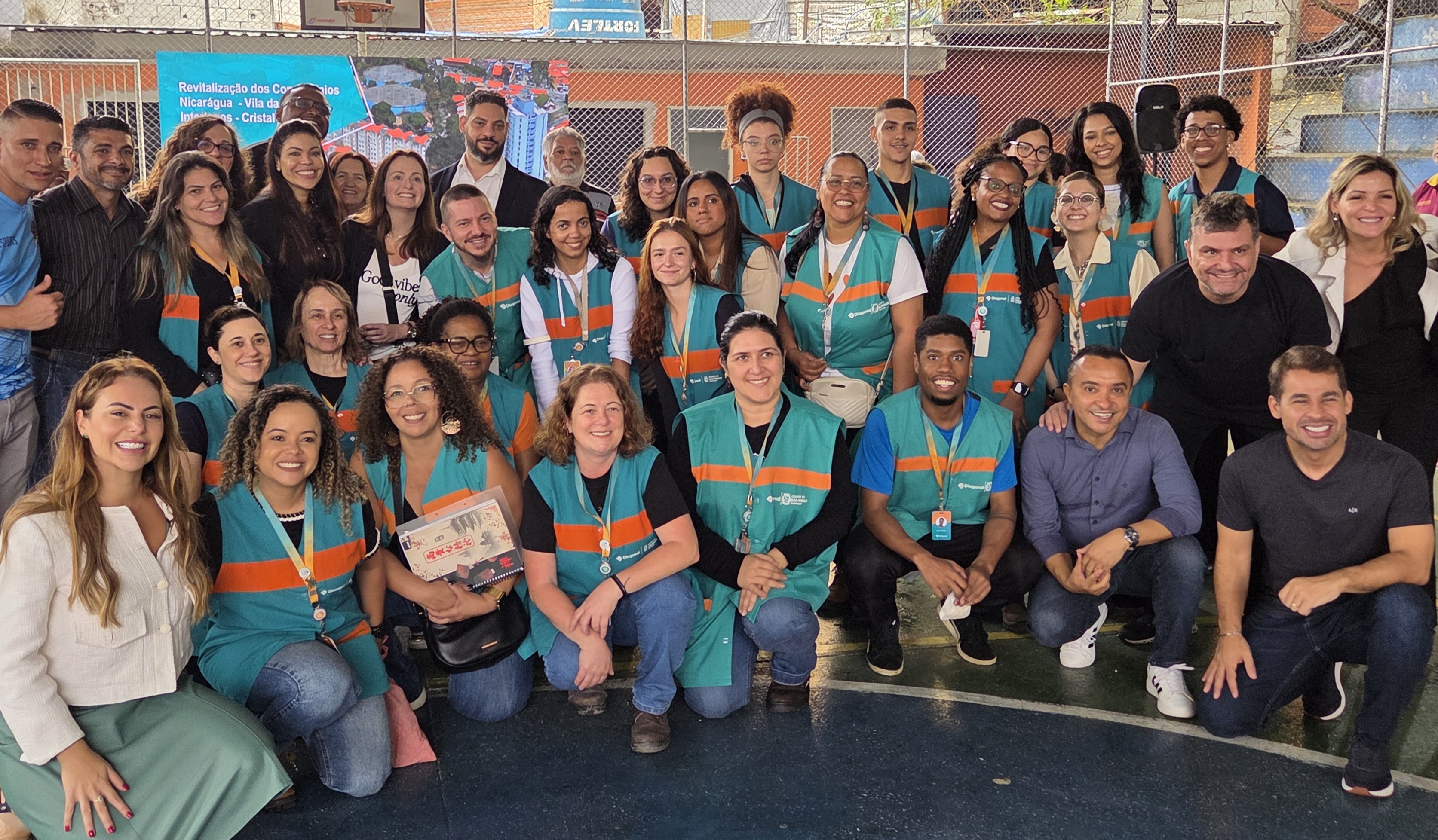 Secretário Sidney Cruz junto com Diogo Soares, Cacá Viana e Regina Nunes junto com a equipe de trabalho social reunidos em uma quadra coberta para foto coletiva. A maioria veste coletes verdes com faixas laranja, e todos sorriem para a câmera. 