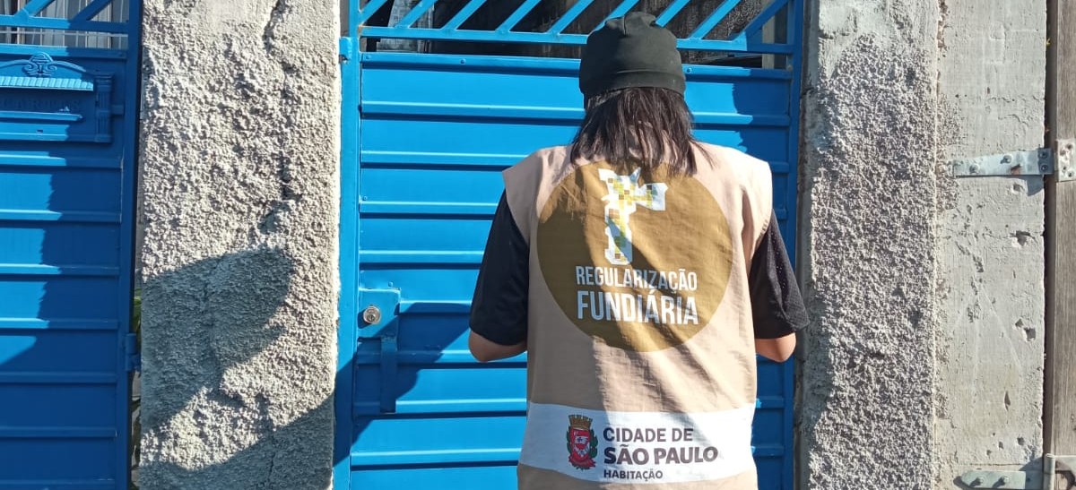 Mulher de costas realizando o cadastramento de uma casa. Ela veste o colete bege com a logotipo da Regularização Fundiária, escrito 