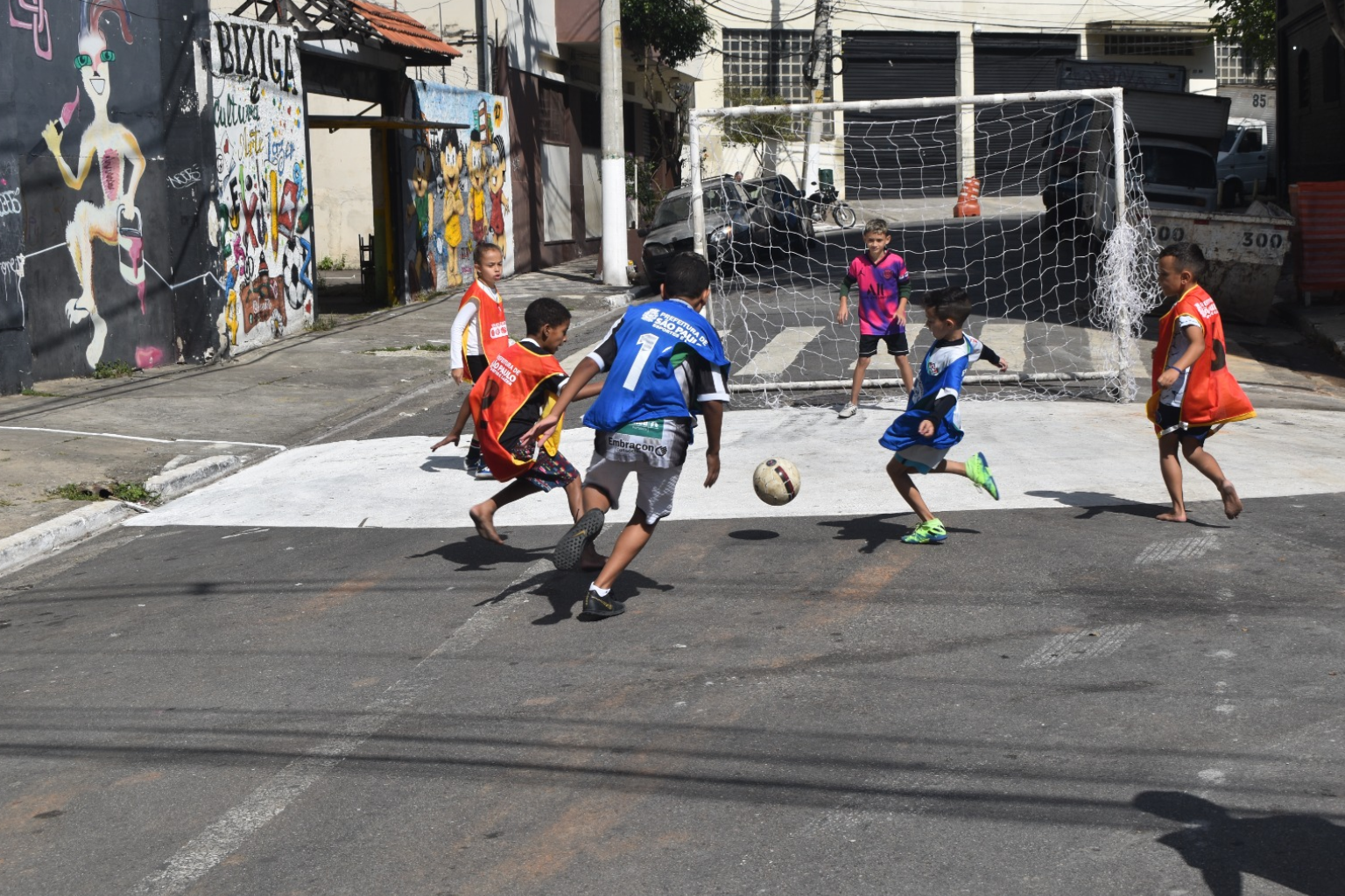 Imagem de crianças com coletes azuis e laranjas jogando futebol em um rua pública de lazer 