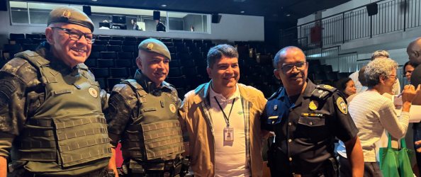 A imagem mostra quatro homens posando para uma foto em um ambiente interno, que parece ser um auditório. Três deles estão vestidos com uniformes militares ou policiais: dois usam fardas camufladas com coletes à prova de balas e boinas verdes, e um está com uniforme escuro da Guarda Civil Metropolitana (GCM), com distintivos visíveis. Entre eles, o Subprefeito Guilherme Henriques, vestindo uma jaqueta bege clara, camisa branca e crachá no pescoço, sorrindo para a foto. Ao fundo, há várias cadeiras vazias e algumas pessoas conversando em pé, indicando um evento ou cerimônia pública. O ambiente é iluminado artificialmente e transmite uma atmosfera institucional e cordial.