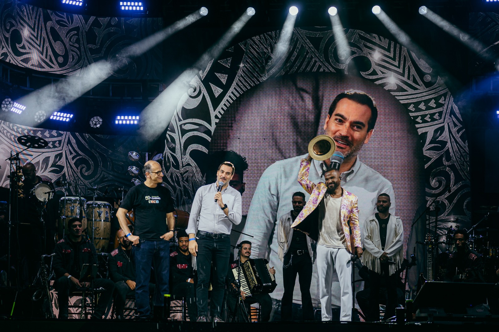 escrição geral: Fotografia de palco tirada durante um evento noturno, mostrando três figuras em destaque perante o público, com músicos e um ecrã gigante ao fundo.   Elemento central: No palco, encontram-se três homens a falar ou a interagir: à esquerda, um homem de óculos e camisa escura; ao centro, o secretário a segurar um microfone; e à direita, o cantor Alexandre Pires vestido com um casaco estampado e calças brancas, a segurar um trompete.   Músicos e cenário: Ao fundo, vários membros da banda estão posicionados junto a instrumentos musicais (incluindo acordeão e percussão), em frente a um grande ecrã que projeta o rosto de um dos intervenientes.   Iluminação: O cenário é iluminado por fortes focos de luz vertical que criam feixes brancos brilhantes no topo do palco escuro.   