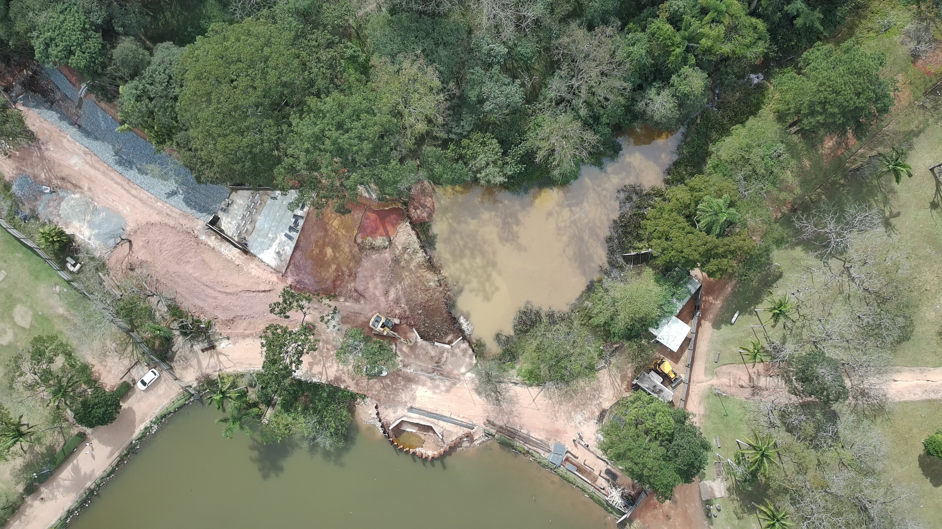Fotografia aérea em vista superior (planta baixa) mostrando o canteiro de obras de drenagem situado entre dois corpos d'água no Parque do Carmo, em Itaquera.  No centro da imagem, há uma faixa de terra avermelhada e mexida que conecta as duas áreas aquáticas. Sobre esse caminho de terra, encontra-se uma escavadeira amarela em operação próxima à margem do lago superior.  Na parte inferior da foto, vê-se um trecho de um grande lago de águas esverdeadas, com estruturas de drenagem de concreto e estacas-pranchas metálicas instaladas na borda. Na parte central e superior direita, há um segundo corpo d'água menor, com águas turvas de tom marrom-amarelado.  Ao redor de todo o canteiro de obras e das áreas de água, há extensa vegetação com árvores de copas verdes e densas, gramados e caminhos de terra batida. No canto inferior esquerdo, observa-se um veículo branco estacionado próximo a uma das vias internas do parque.
