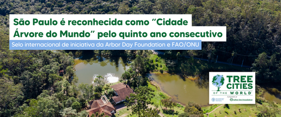 Esta imagem apresenta uma vista aérea de uma área de preservação com mata densa e um lago central sob a luz do sol. No canto superior esquerdo, um texto em verde e branco destaca que a cidade de São Paulo foi reconhecida como 