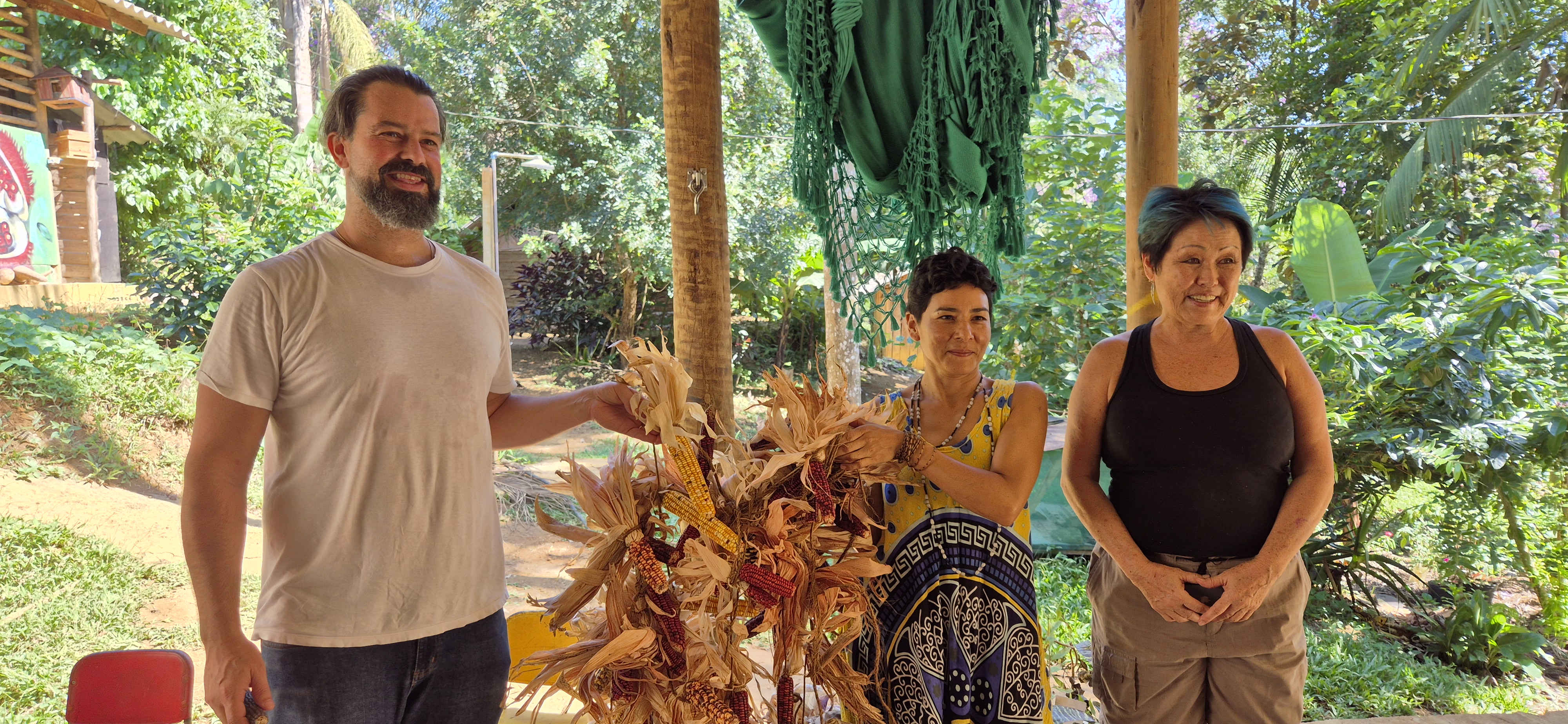 Imagem de três pessoas em aldeia guarani posando para foto. Há um homem do lado esquerdo, que ajuda uma mulher que está no centro a segurar espigas de milho. A outra mulher no canto direito posa para foto. Todos estão sorrindo. No fundo há várias árvores, indicando que estão em uma área verde. 