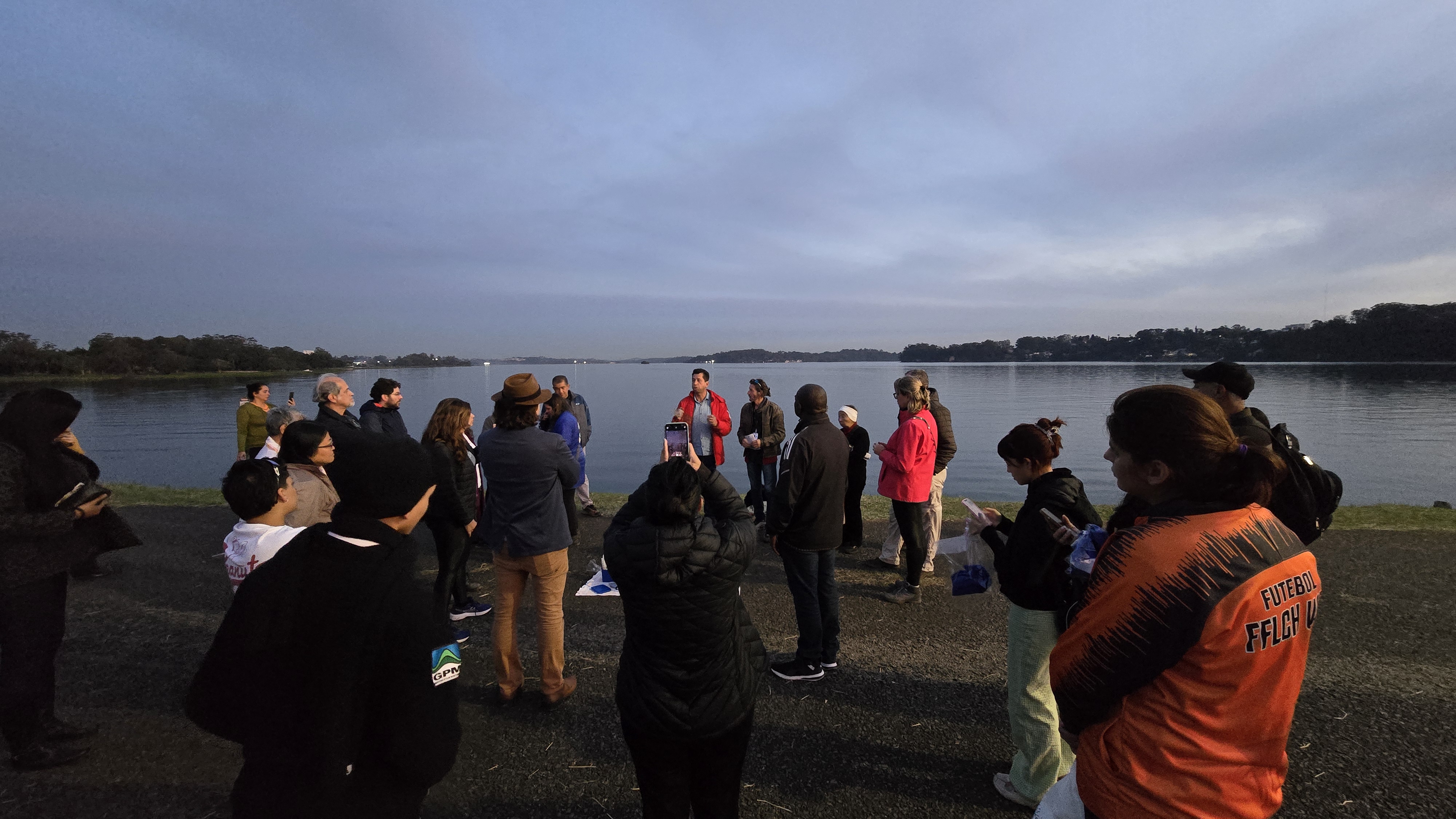 Grupo de cerca de 25 pessoas reunido ao ar livre, em roda, próximo à margem de um grande lago ao entardecer. No centro, um homem de jaqueta vermelha fala ao grupo, enquanto alguns participantes observam, tiram fotos ou seguram anotações. O céu está nublado, com tons azulados, e a água do lago reflete a paisagem tranquila. A cena transmite um momento de conversa ou orientação durante uma atividade coletiva em ambiente natural.