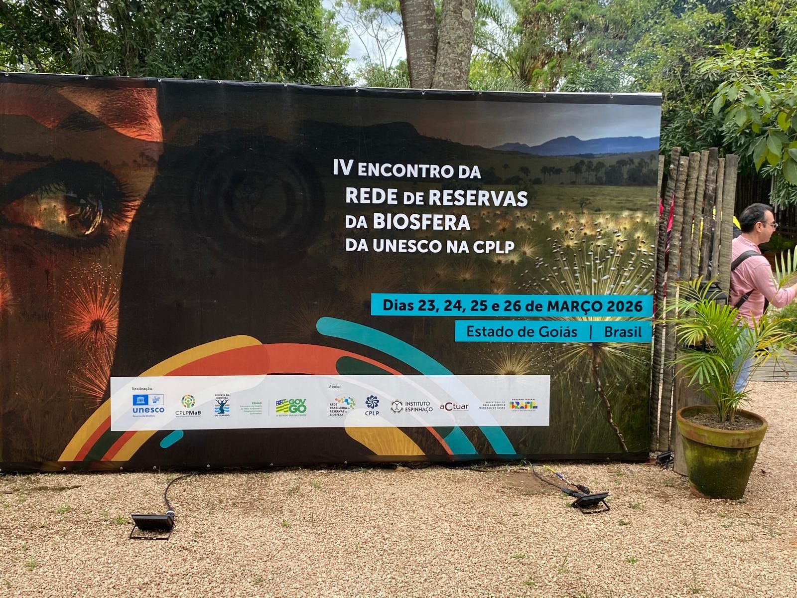 A imagem mostra um ambiente interno com estrutura rústica, com teto de madeira em tons claros e vigas em marrom escuro, iluminado por luz natural que entra pelas laterais, onde há vegetação ao fundo; no centro, um grande telão branco exibe o brasão de Angola em azul, amarelo e vermelho, acompanhado de textos em preto e cinza, com destaque para o título “Reserva da Biosfera da Quiçama Brasil-Março-2026”, além de logotipos coloridos em verde, azul e laranja na parte inferior; à direita, uma mulher em pé, vestindo roupa em tom bege claro ou cinza, apresenta o conteúdo, tendo atrás de si um banner escuro com textos em branco e verde; em primeiro plano, várias pessoas estão sentadas em mesas de madeira em tons quentes de marrom, utilizando roupas claras e escuras, enquanto um homem à esquerda segura um celular para registrar a cena, e sobre as mesas há copos, garrafas e notebooks, incluindo um aberto com tela escura refletindo o ambiente; ao centro inferior, um arranjo de plantas secas em tons naturais — bege, marrom e rosado — complementa a composição, cuja paleta mistura cores neutras com pontos de cor mais vivos vindos do telão e dos elementos gráficos.
