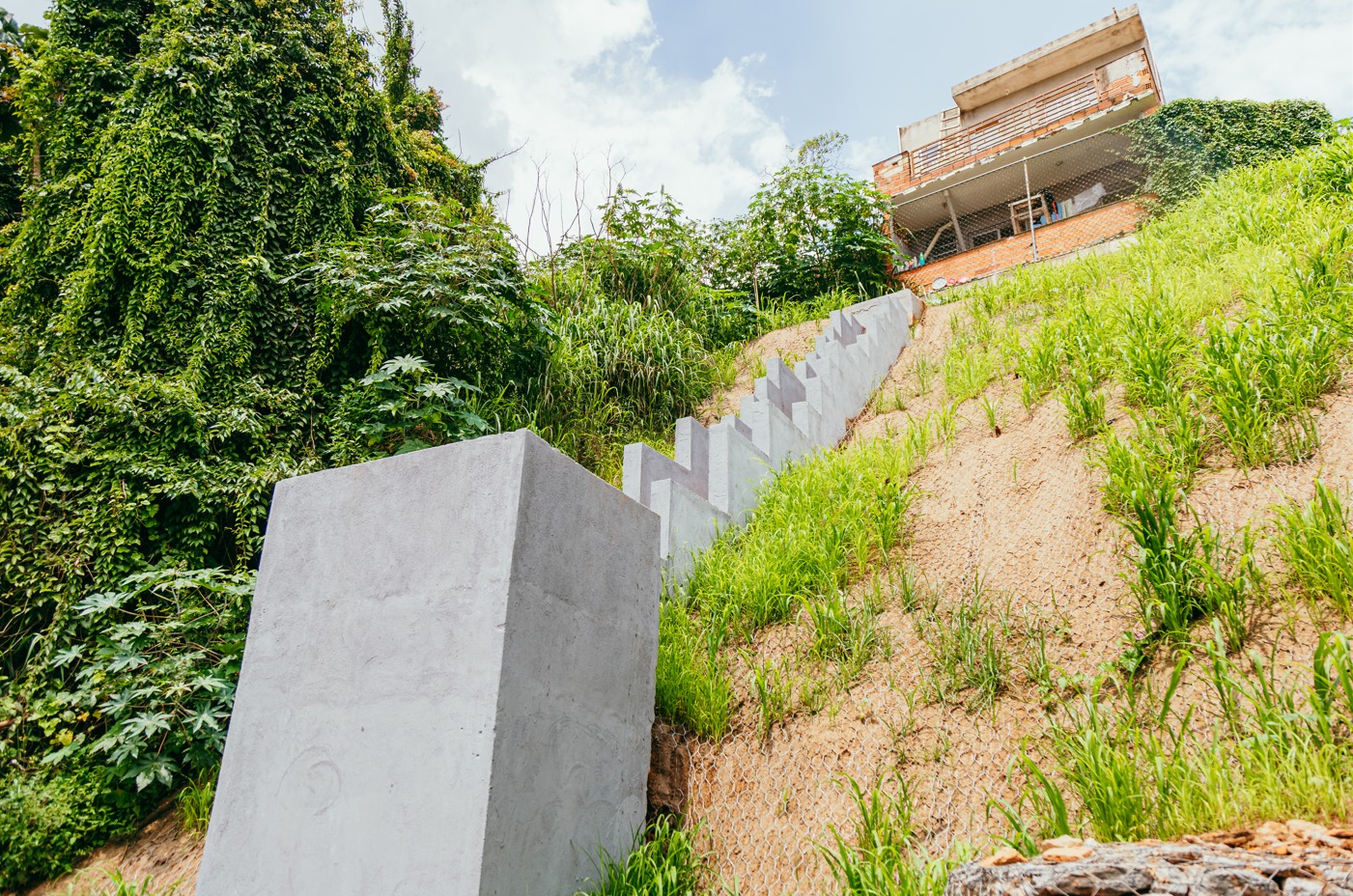 Imagem aérea apresenta um conjunto arquitetônico com prédio térreo de tijolos aparentes e cobertura vegetal. Em frente à construção, há uma área externa com arquibancadas em concreto dispostas em níveis, intercaladas por faixas de grama. O espaço conta com piso intertravado, postes de iluminação, pequenas árvores recém-plantadas e equipamentos ao ar livre. Ao fundo, observa-se área verde cercada e vias urbanas próximas, indicando equipamento público inserido em contexto urbano.