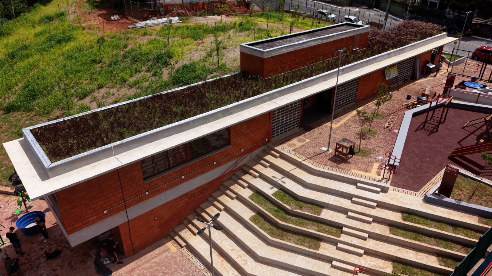 Imagem mostra um talude íngreme coberto por vegetação, com gramíneas e trepadeiras já estabelecidas sobre o solo. No centro do morro há uma estrutura de contenção em concreto, composta por placas alinhadas de forma escalonada, que contribuem para a estabilização da encosta. No topo do terreno, observa-se uma casa de alvenaria com varanda e guarda-corpo metálico. O cenário evidencia uma intervenção de engenharia voltada à prevenção de erosão em área residencial.
