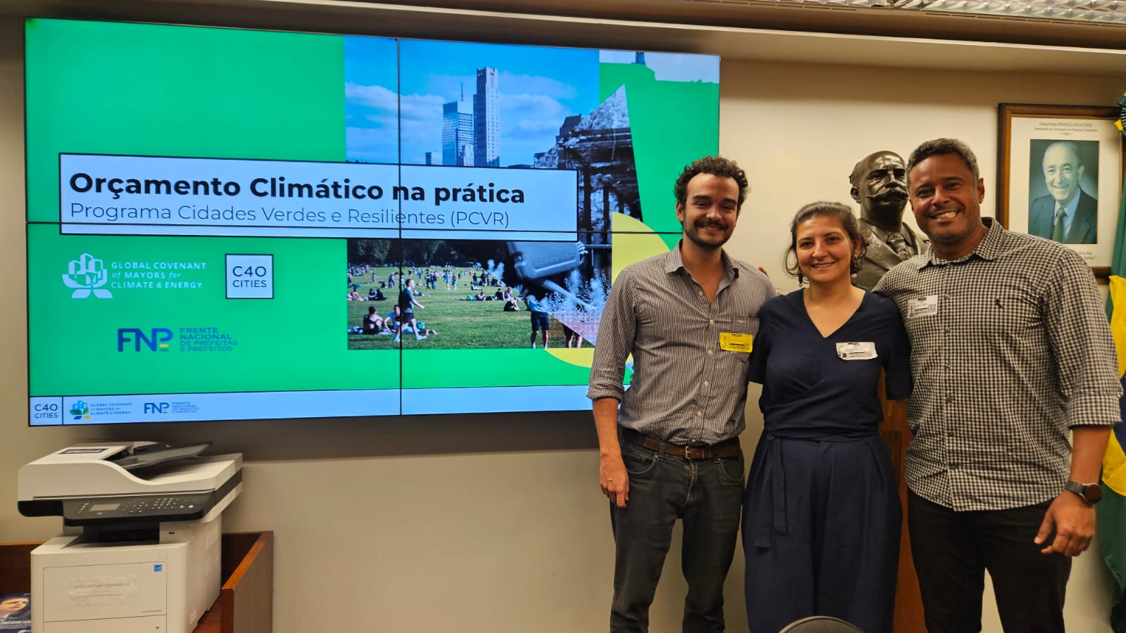 **Para cego ver:** Três pessoas posam sorrindo em um ambiente institucional ao lado de um grande painel digital. Na tela aparece o título “Orçamento Climático na prática – Programa Cidades Verdes e Resilientes (PCVR)”, acompanhado de logos de organizações ligadas à agenda climática. Ao fundo, há um busto escultórico e quadros na parede. O grupo está em um espaço de reunião ou evento voltado a políticas públicas e sustentabilidade.