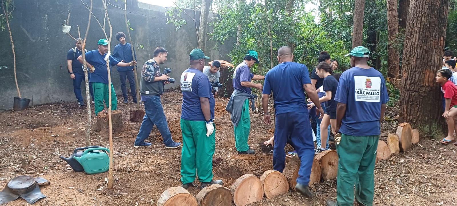 A imagem mostra um grupo de pessoas participando de uma atividade coletiva em meio a uma área arborizada, com destaque para o plantio de mudas. Os uniformes azul-marinho com verde, usados por alguns participantes, contrastam com o marrom avermelhado da terra revolvida e os troncos dispostos ao redor. O cenário é emoldurado por tons terrosos do solo e dos troncos de árvores, além dos verdes vivos da vegetação ao fundo. As cores naturais predominam, reforçando o ambiente de trabalho ambiental, com a presença de agentes da Prefeitura de São Paulo e outros colaboradores envolvidos na ação ecológica.