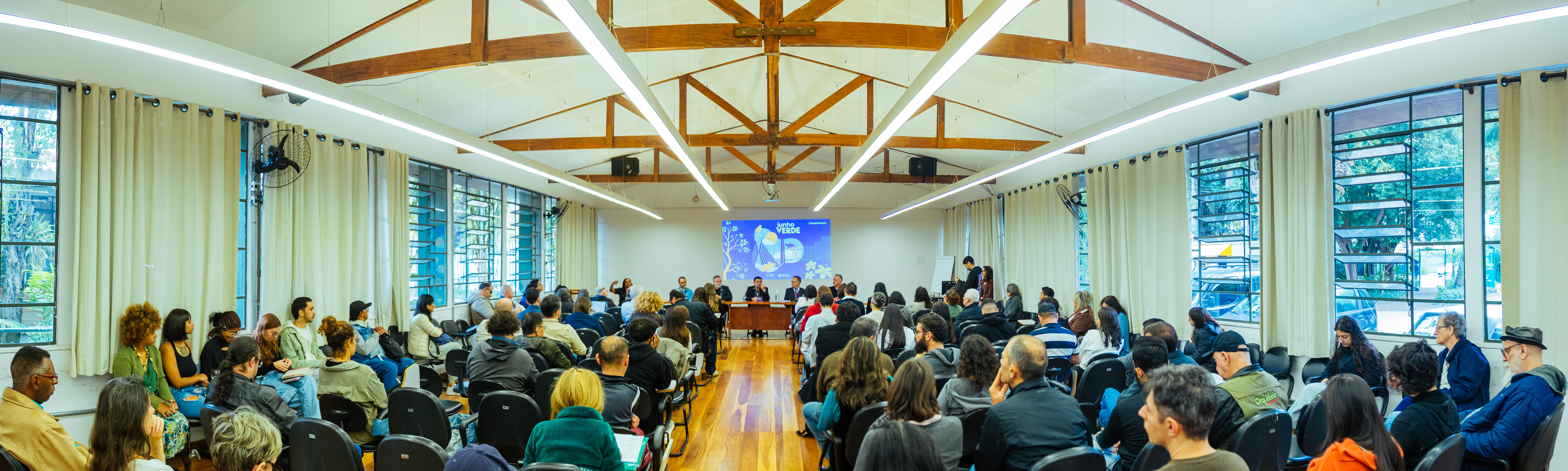 Sala ampla e bem iluminada, com estrutura de madeira no teto e grandes janelas com cortinas claras nas laterais. A imagem mostra um auditório cheio, com dezenas de pessoas sentadas em cadeiras organizadas em fileiras, assistindo a uma mesa de debate localizada à frente da sala. Ao fundo, uma tela exibe o título “Junho Verde”. O público é diverso, com presença de pessoas de diferentes idades e perfis, demonstrando atenção aos palestrantes. O ambiente remete a um evento institucional ou seminário com foco em temas ambientais.