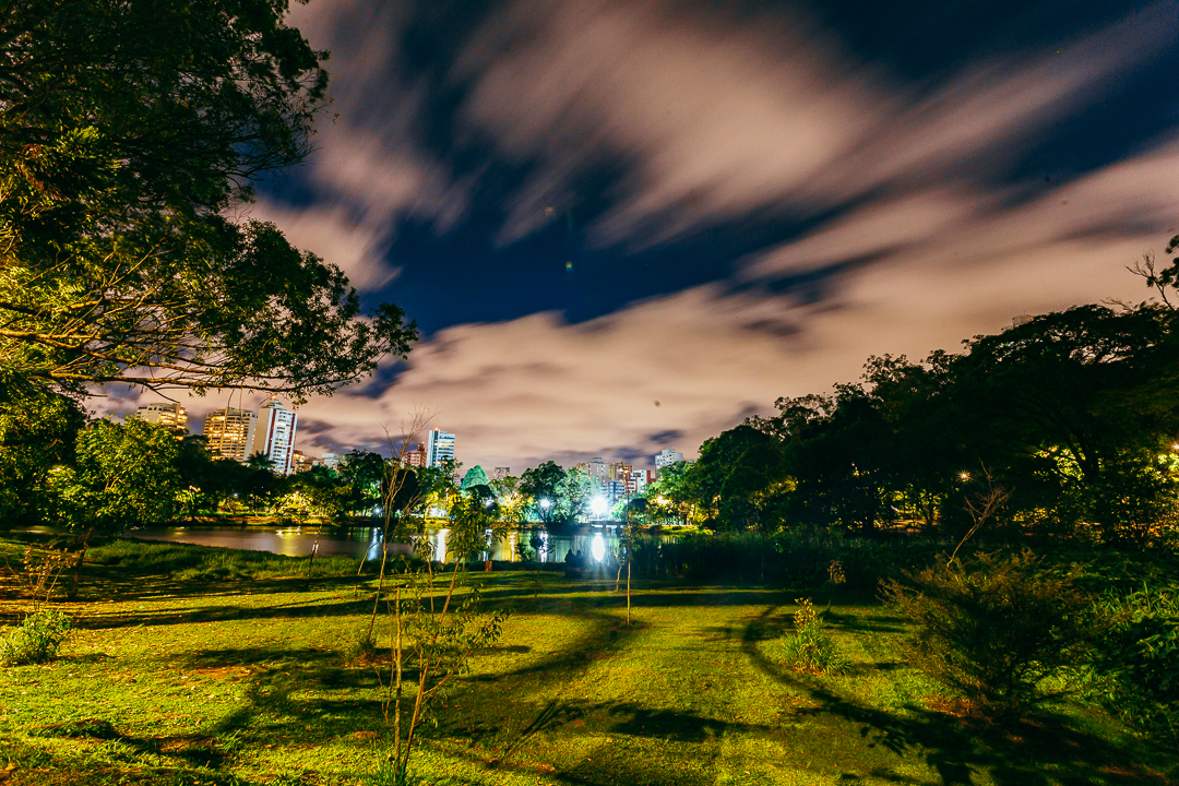 Vista noturna do Parque da Aclimação, em São Paulo, com céu azul-escuro e nuvens em movimento. A iluminação artificial destaca a vegetação e o gramado do parque, enquanto o espelho d’água reflete as luzes dos prédios ao fundo. Árvores emolduram a imagem dos dois lados e os edifícios iluminados ao fundo contrastam com a natureza em primeiro plano.