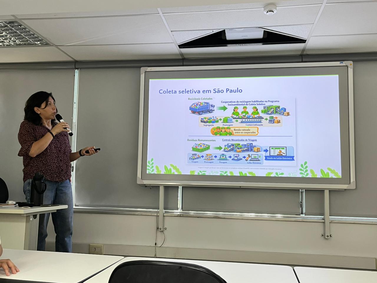 A imagem mostra uma sala de aula ou auditório com iluminação clara e tons neutros. À esquerda, há uma mulher em pé fazendo uma apresentação. Ela tem cabelo escuro preso, veste uma blusa de manga curta com estampa em tons de vinho e branco e calça jeans azul. Na mão direita, segura um microfone preto, e na esquerda, um controle para passar slides. Sua expressão é atenta e focada, olhando em direção à tela.  Ao fundo, há uma tela de projeção branca exibindo um slide com o título “Coleta seletiva em São Paulo”, escrito em azul. O slide contém ilustrações coloridas, com predominância de verde, azul e amarelo, mostrando etapas da coleta e reciclagem de resíduos, como caminhões, pessoas trabalhando e materiais recicláveis. Na parte inferior do slide, há detalhes em verde que lembram folhas ou elementos naturais.  O ambiente tem paredes claras e um teto branco com placas modulares. Uma parte do teto está aberta, revelando um equipamento interno. Na frente da sala, há mesas brancas e cadeiras pretas. No canto inferior esquerdo da imagem, aparece parcialmente o braço de outra pessoa sentada, indicando a presença de público.