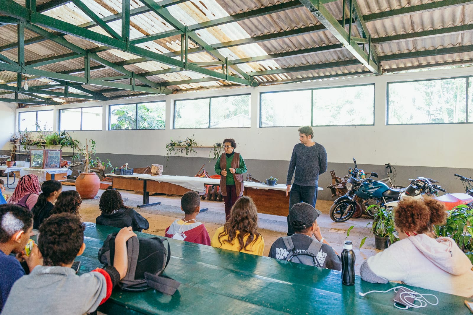 Fotografia de uma atividade de educação ambiental do programa Aventura Ambiental, realizada em um espaço amplo e coberto. Ao fundo, há janelas, mesas, plantas e motocicletas. No centro da imagem, uma mulher está em pé diante de um grupo de participantes, que estão sentados em mesas e voltados para ela; ao lado dela, há um homem também em pé. A mulher parece conduzir a atividade enquanto o grupo acompanha a apresentação. Na parte inferior da imagem, aparecem principalmente as pessoas sentadas, vistas de costas.