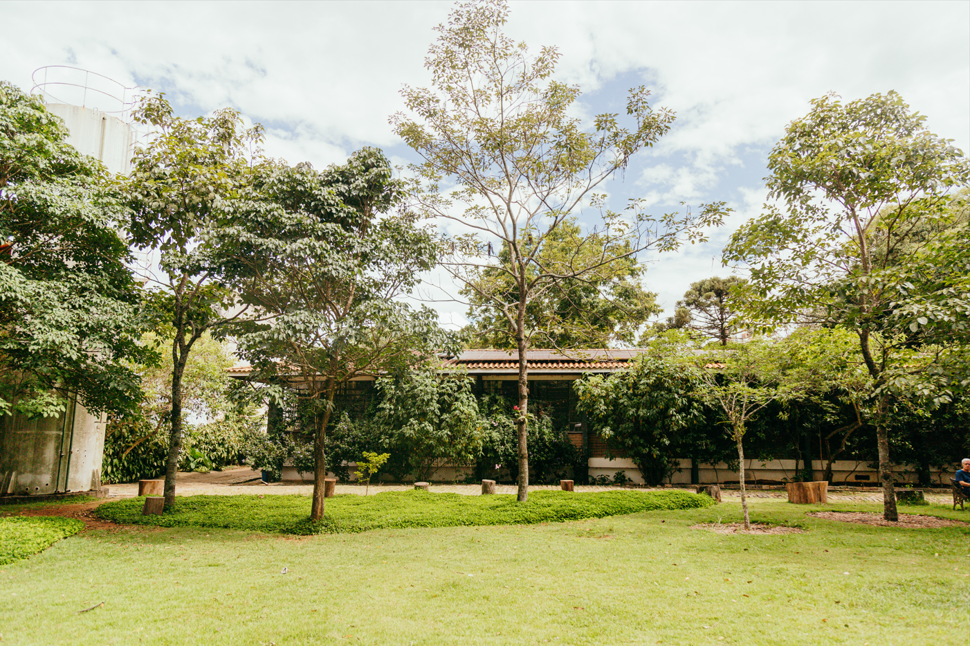 A imagem mostra um amplo jardim com gramado bem cuidado e diversas árvores jovens e adultas espalhadas de maneira harmoniosa pelo espaço. Ao fundo, parcialmente encoberta pela vegetação, há uma construção térrea com telhado de telhas cerâmicas e paredes de alvenaria clara com detalhes em tijolos aparentes. A fachada da edificação é sombreada por arbustos e trepadeiras, que criam uma integração visual com o entorno verde. À esquerda da imagem, vê-se um grande reservatório cilíndrico de concreto, sugerindo a presença de infraestrutura para captação ou armazenamento de água. No primeiro plano, há tocos de madeira distribuídos como bancos rústicos em áreas de convivência sob as árvores, reforçando o caráter natural e acolhedor do local. O céu parcialmente nublado e a iluminação suave indicam um dia claro, com boa incidência de luz natural. Um homem sentado à direita da imagem, próximo a um dos troncos, completa a cena tranquila e arborizada.