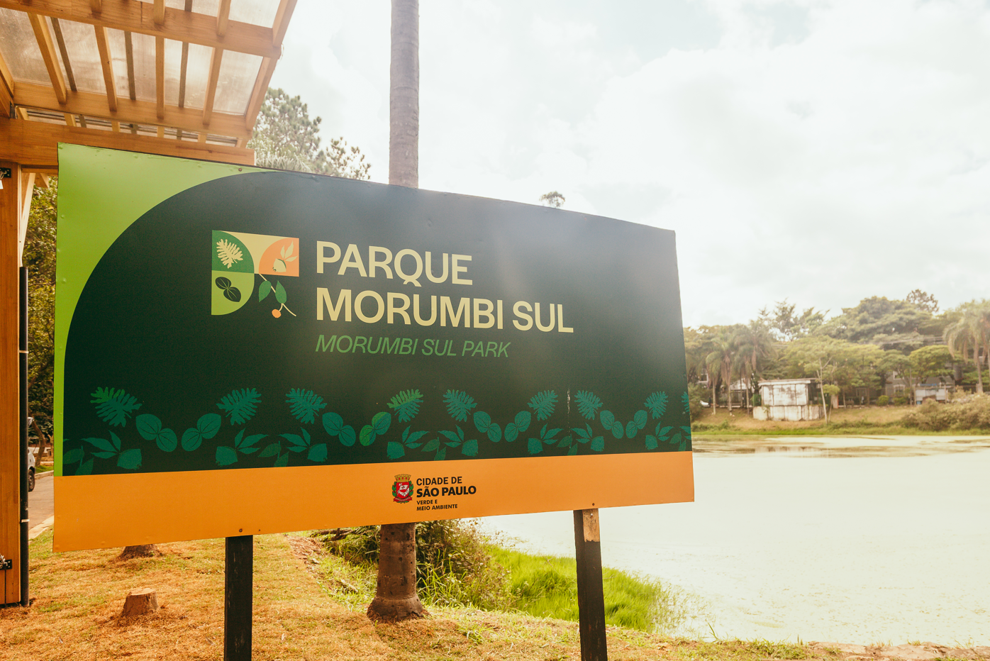 A imagem mostra a placa de identificação do Parque Morumbi Sul, localizado na cidade de São Paulo. A placa tem design moderno e utiliza cores verde-escuras com detalhes em laranja e verde-claro, além de ilustrações botânicas que remetem à flora local. Abaixo do nome do parque, há a tradução em inglês: "Morumbi Sul Park", indicando acessibilidade para visitantes estrangeiros.  Na parte inferior da placa está o selo da Cidade de São Paulo – Verde e Meio Ambiente, sugerindo que o parque é administrado pela Secretaria Municipal do Verde e do Meio Ambiente.  Ao fundo, é possível ver um corpo d’água e, mais adiante, vegetação densa e edificações residenciais. A presença de lago, árvores e estrutura de madeira ao lado da placa reforça o perfil de parque urbano com proposta ecológica, de lazer e recuperação ambiental — características alinhadas às diretrizes de sustentabilidade da cidade.