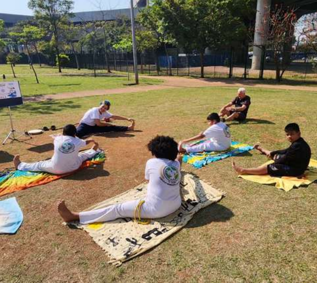 A foto mostra uma atividade ao ar livre em um parque, onde um grupo de pessoas está sentado em círculo sobre toalhas ou esteiras, realizando alongamentos.  A maioria veste roupas brancas, típicas de uniformes de capoeira, com cordas coloridas na cintura. Isso indica que provavelmente se trata de uma aula ou oficina de capoeira, começando com exercícios de preparação física.  O ambiente é arborizado, com gramado, árvores ao fundo e uma pista de caminhada passando atrás do grupo, transmitindo um clima de prática comunitária, saudável e em contato com a natureza.