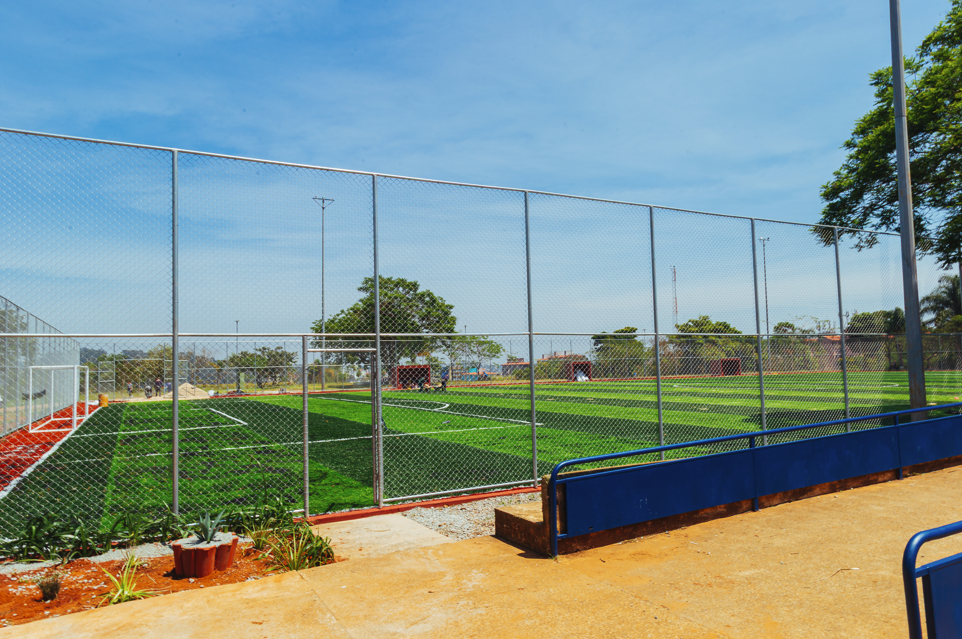 Imagem de um campo de futebol com grama sintética em tons de verde, formando faixas mais claras e mais escuras. O campo é cercado por uma tela metálica prateada. No primeiro plano, há um piso em tom bege/amarelado e, à direita, uma arquibancada baixa azul. À esquerda, aparece parte da área do gol com estrutura branca e detalhes em vermelho no entorno. Próximo ao chão, há pequenos canteiros com plantas verdes e terra avermelhada. Ao fundo, vê-se árvores verdes, algumas construções em cores neutras e um céu azul claro, indicando um dia ensolarado.