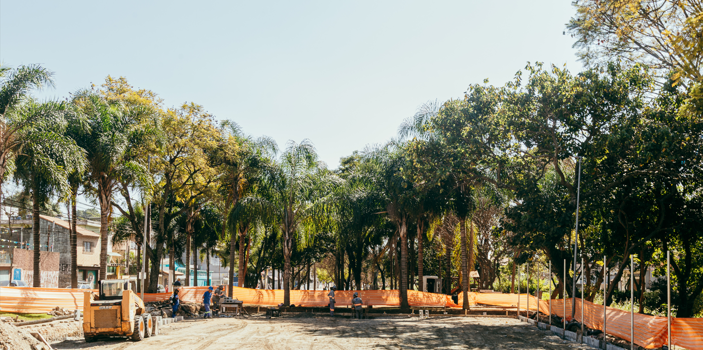 Imagem mostra uma área urbana em processo de requalificação. Em primeiro plano, há um terreno amplo de terra batida, com máquinas de obra e alguns trabalhadores utilizando equipamentos. O espaço está parcialmente cercado por telas de proteção laranjas. Ao fundo e nas laterais, há diversas árvores altas, incluindo palmeiras, formando uma área verde. Também é possível ver casas ao redor, indicando que o local fica em um bairro residencial. O céu está claro, com bastante luz natural.