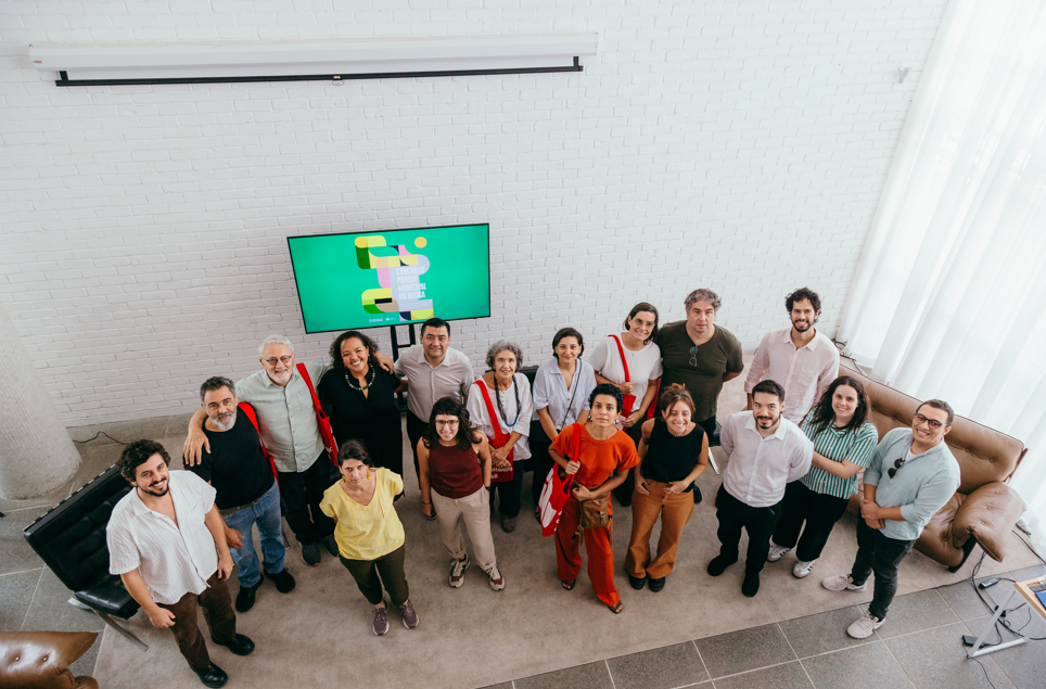 Foto tirada de cima mostra um grupo de cerca de 18 pessoas reunidas em um espaço interno bem iluminado. Elas estão próximas umas das outras, olhando para a câmera e sorrindo. O ambiente tem parede de tijolos brancos, um sofá marrom à direita e uma TV ao fundo exibindo uma imagem com fundo verde. As pessoas usam roupas casuais, com cores variadas, e algumas carregam bolsas. O clima é descontraído e coletivo.
