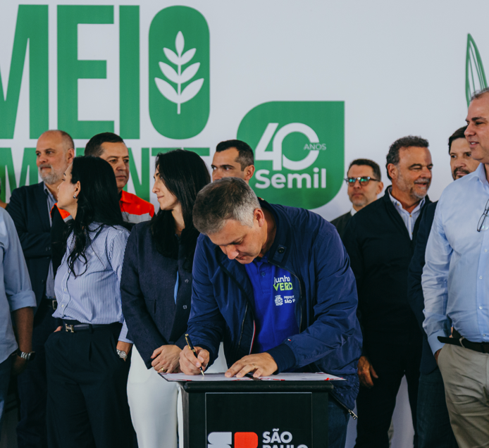 Fotografia de um evento institucional sobre meio ambiente. Em primeiro plano, um homem assina um documento em um púlpito com a marca da Prefeitura de São Paulo. Ao fundo, autoridades e participantes acompanham a cerimônia. No painel de fundo aparecem as palavras “Meio Ambiente” em destaque e o logotipo comemorativo dos 40 anos da Semil (Secretaria de Meio Ambiente, Infraestrutura e Logística do Estado de São Paulo). A imagem registra a assinatura de um termo de cooperação para fortalecer ações de educação ambiental na capital paulista.