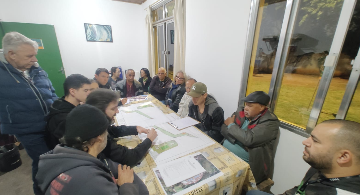 Reunião em uma sala, com cerca de 12 pessoas sentadas ao redor de uma mesa. Sobre a mesa há grandes plantas e mapas do parque, além de documentos. Algumas pessoas analisam os materiais e apontam para os mapas, enquanto as demais acompanham a discussão. Ao fundo, há janelas de vidro com vista para uma área gramada e arborizada.
