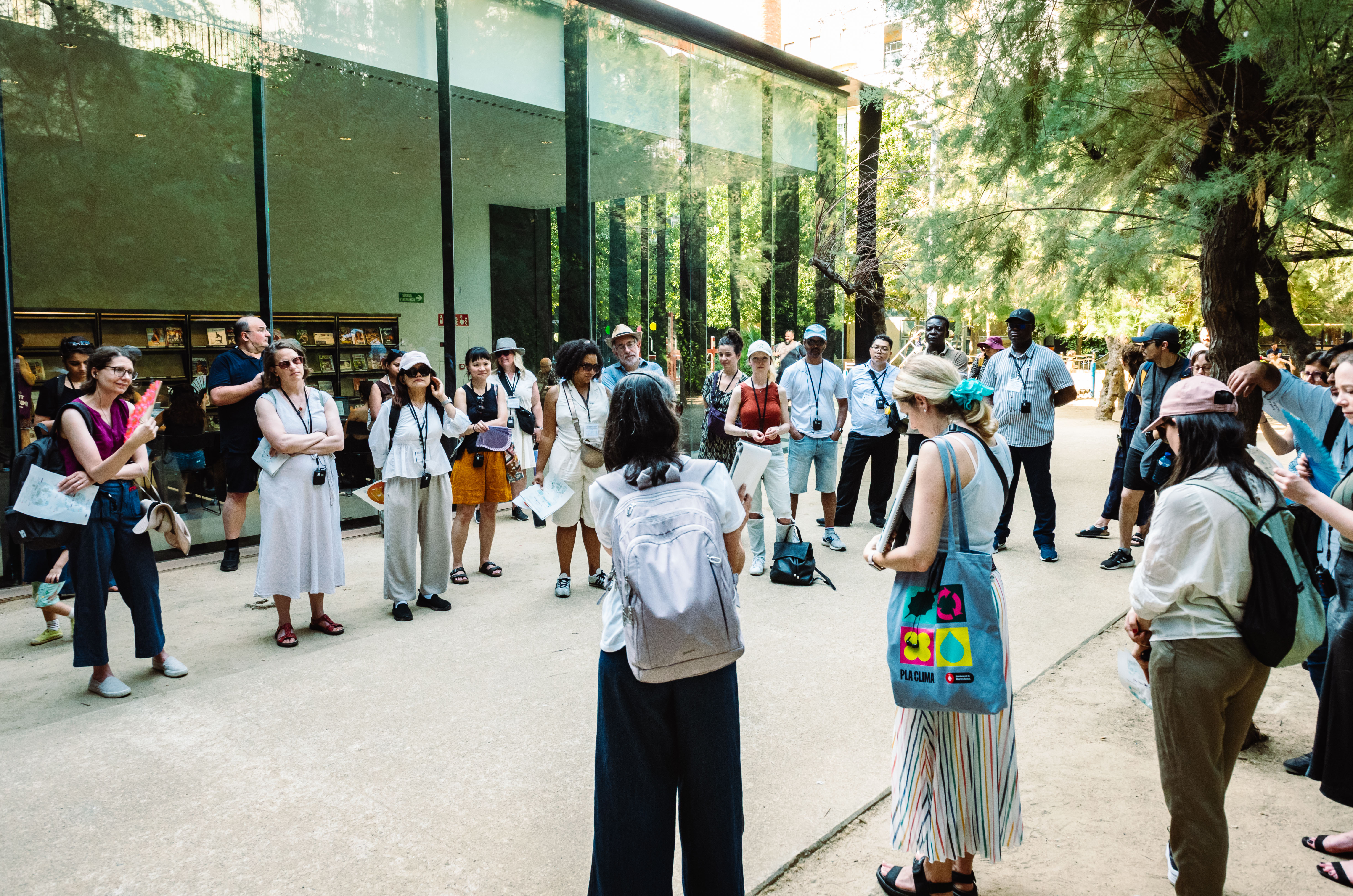 Grupo de pessoas reunido em círculo ao ar livre, em frente a um edifício moderno com fachada de vidro. A maioria usa crachás, mochilas e equipamentos de áudio, indicando participação em uma visita guiada ou atividade técnica. As pessoas escutam atentamente a uma mulher de costas, que segura papéis e parece conduzir a conversa. Ao fundo, árvores e prateleiras com publicações são visíveis através do vidro.