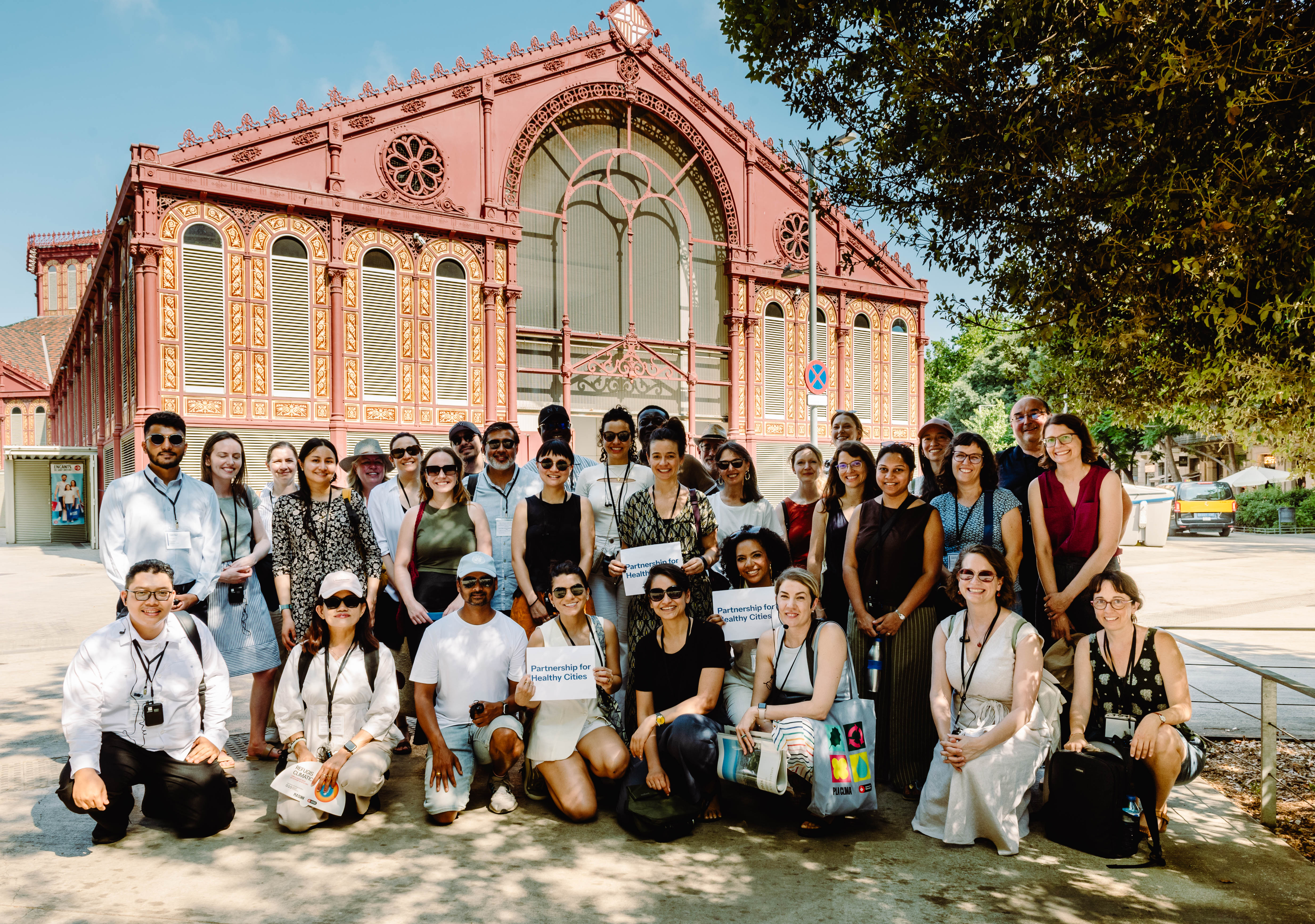 Grupo diverso de cerca de 30 pessoas posa sorridente para foto em frente a um prédio histórico de arquitetura detalhada, com estrutura em vermelho escuro e amarelo. Algumas pessoas seguram cartazes com o nome "Partnership for Healthy Cities". A maioria usa crachás e roupas leves, sugerindo que participam de um evento ou visita institucional em dia ensolarado. Ao fundo, o céu está azul e há árvores que projetam sombra sobre parte do grupo.