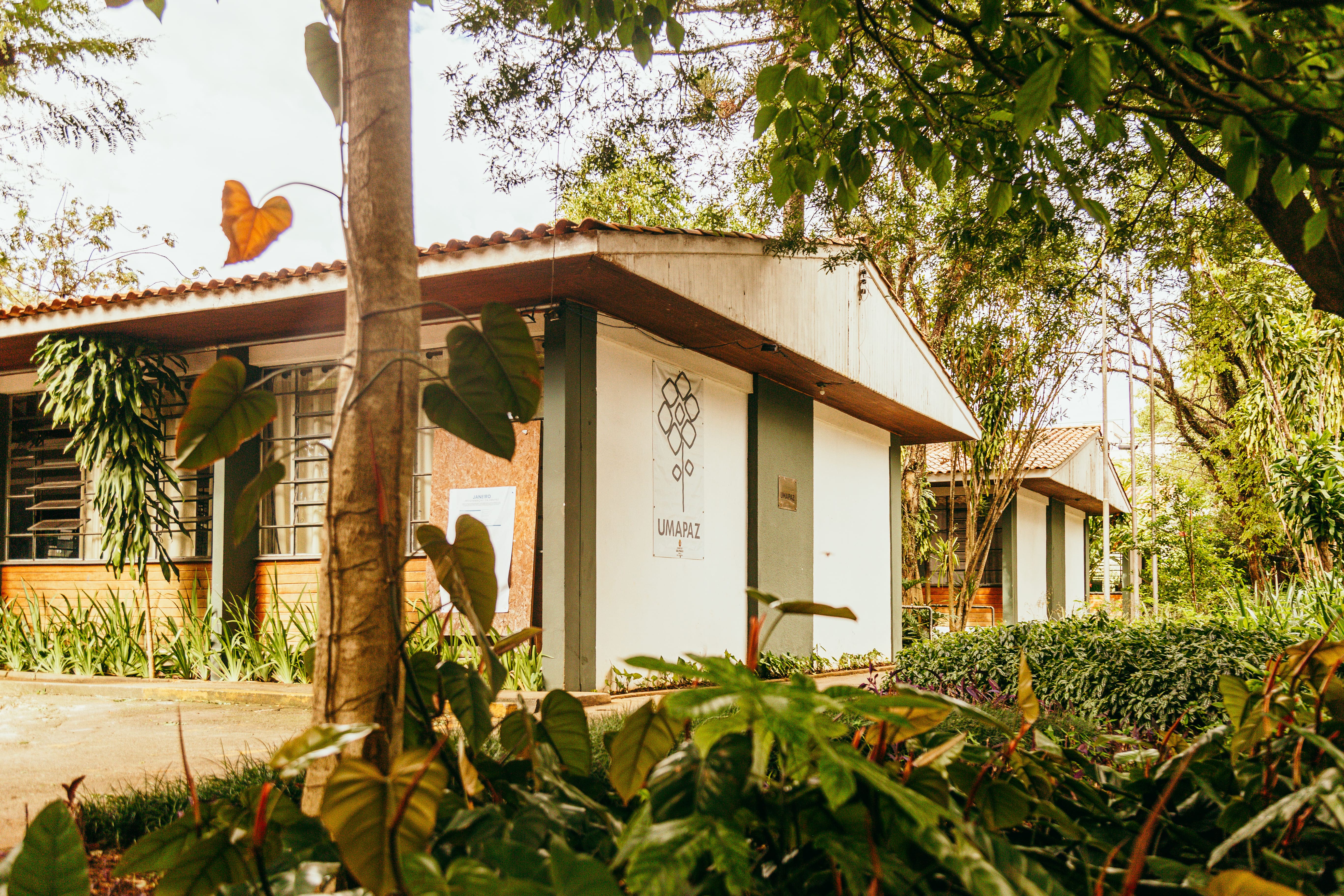 Fachada da sede da UMAPAZ (Universidade Aberta do Meio Ambiente e da Cultura de Paz), localizada em meio à vegetação abundante. O prédio tem estrutura simples, com telhado de cerâmica e paredes brancas com detalhes em verde. No centro da imagem, há uma placa com o logotipo da UMAPAZ fixada na parede. Ao redor, há árvores, plantas ornamentais e folhagens tropicais, compondo um ambiente natural e acolhedor.