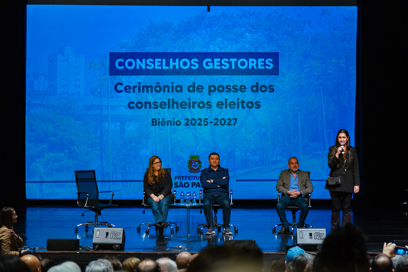 Pessoas participam da cerimônia de posse dos conselheiros eleitos para o biênio 2025-2027 dos Conselhos Gestores. No palco, quatro pessoas estão em destaque: três sentadas em cadeiras giratórias e uma mulher em pé falando ao microfone. Ao fundo, um telão azul projeta o título do evento e o brasão da Prefeitura de São Paulo. O auditório está lotado e parte do público aparece na parte inferior da imagem.