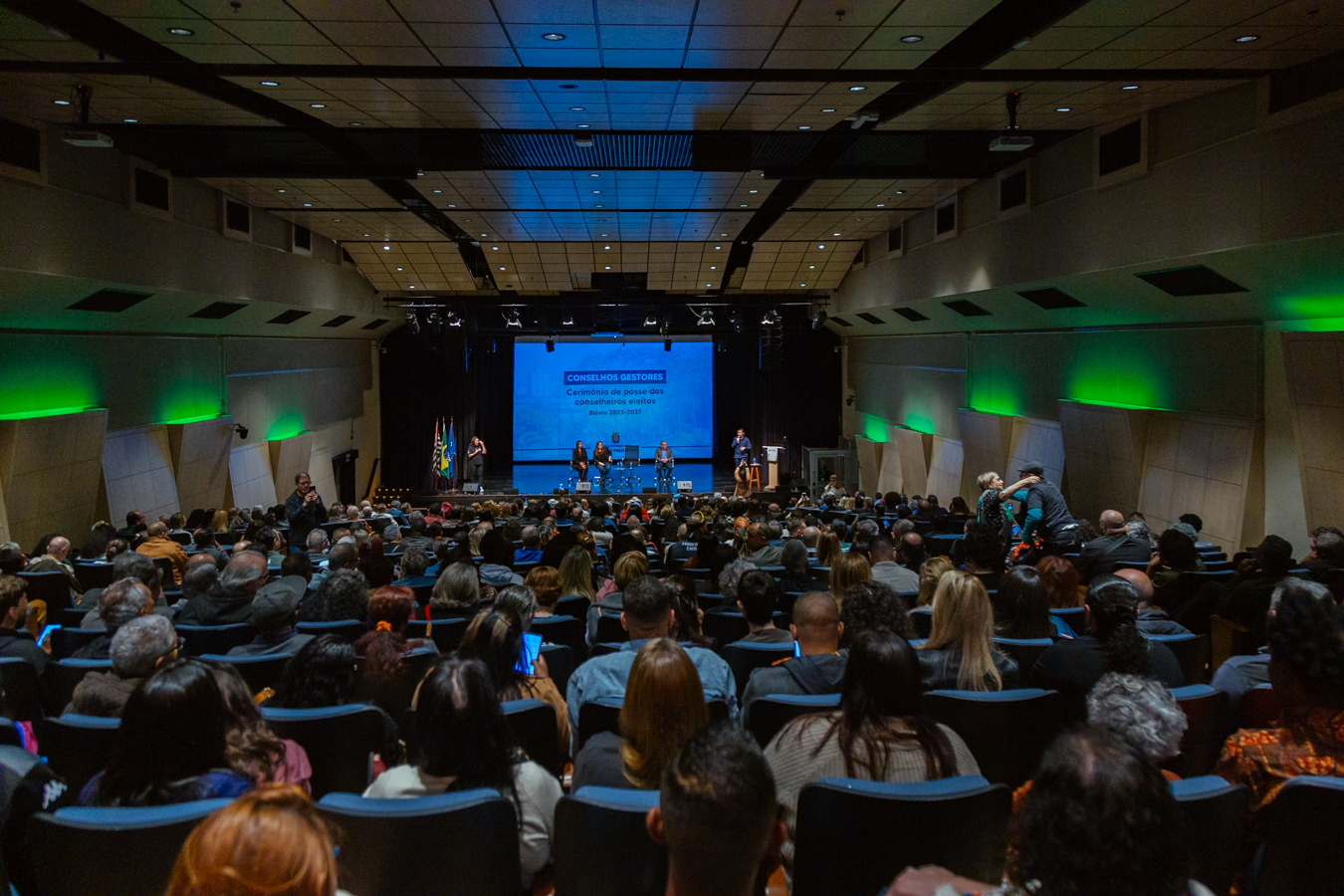 Auditório lotado com dezenas de pessoas acompanhando a cerimônia de posse dos conselheiros eleitos para o biênio 2025-2027 dos Conselhos Gestores. No palco, um telão azul exibe o nome do evento, e algumas pessoas estão sentadas e outras em pé, fazendo pronunciamentos. À esquerda do palco, estão posicionadas bandeiras do Brasil, do estado de São Paulo e da cidade. O ambiente é iluminado com luzes verdes nas paredes laterais.