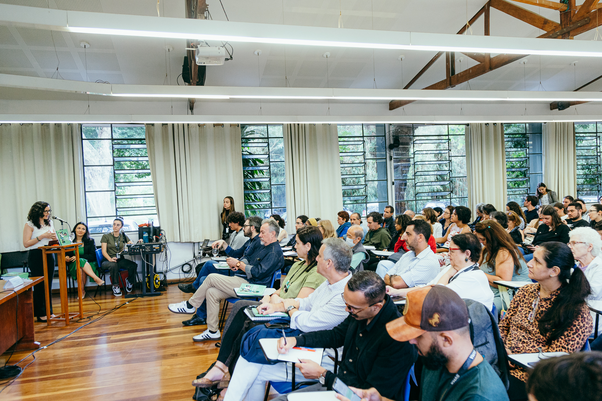 Sala de evento com público sentado em cadeiras, assistindo a uma apresentação. À esquerda, uma mulher fala ao microfone ao lado de uma mesa com computador e equipamentos de som, enquanto outra pessoa está sentada ao seu lado. Os participantes, de diferentes idades, prestam atenção, alguns anotando em cadernos. O espaço é amplo, com janelas grandes ao fundo, cortinas claras e iluminação moderna no teto.