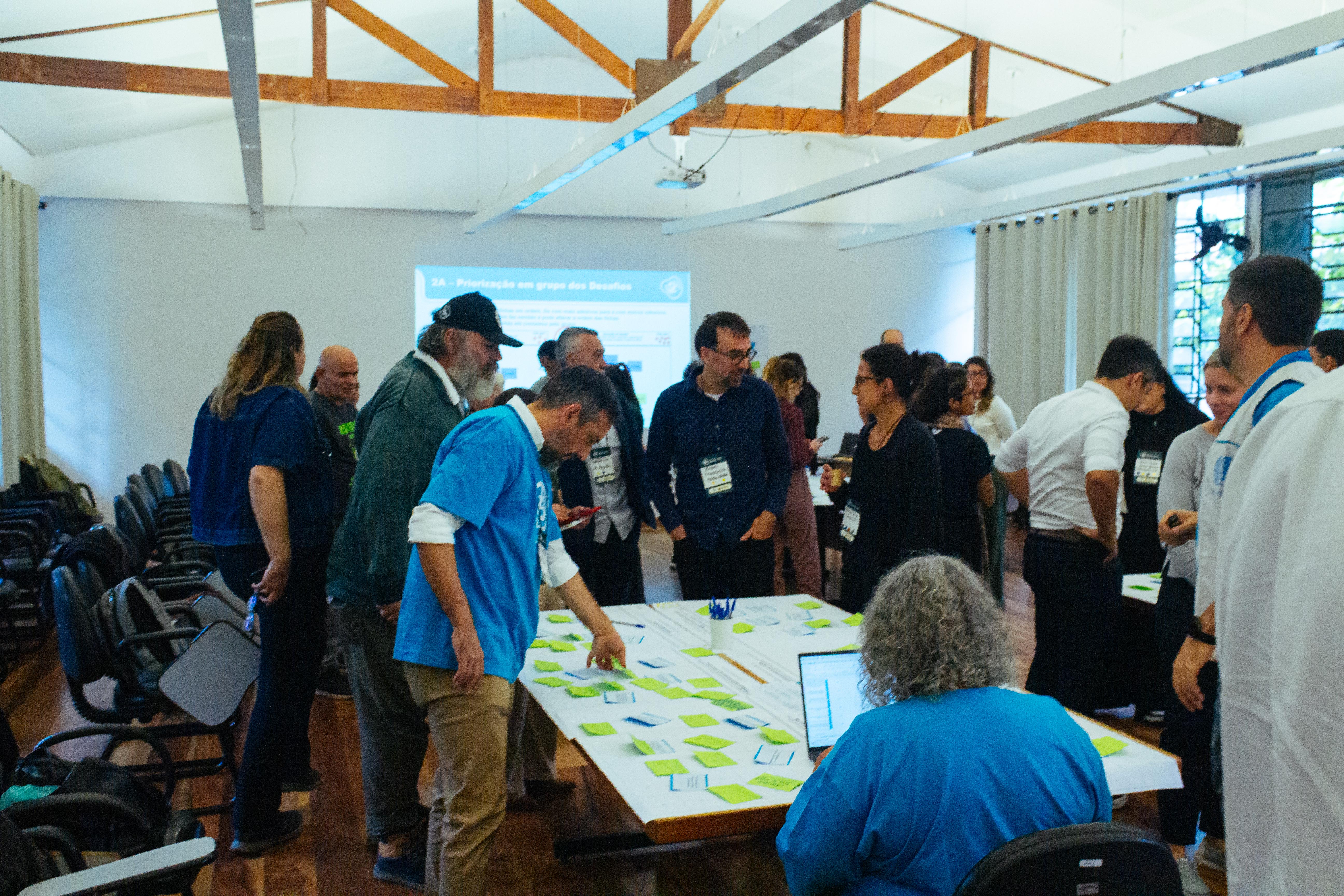Grupo de pessoas participa de uma oficina em uma sala com mesas e cadeiras. No centro, há uma mesa coberta por papéis e post-its coloridos, enquanto alguns participantes analisam e organizam o material. Ao fundo, há uma projeção em tela e outras pessoas em pé conversando, em um ambiente de trabalho colaborativo e educativo.