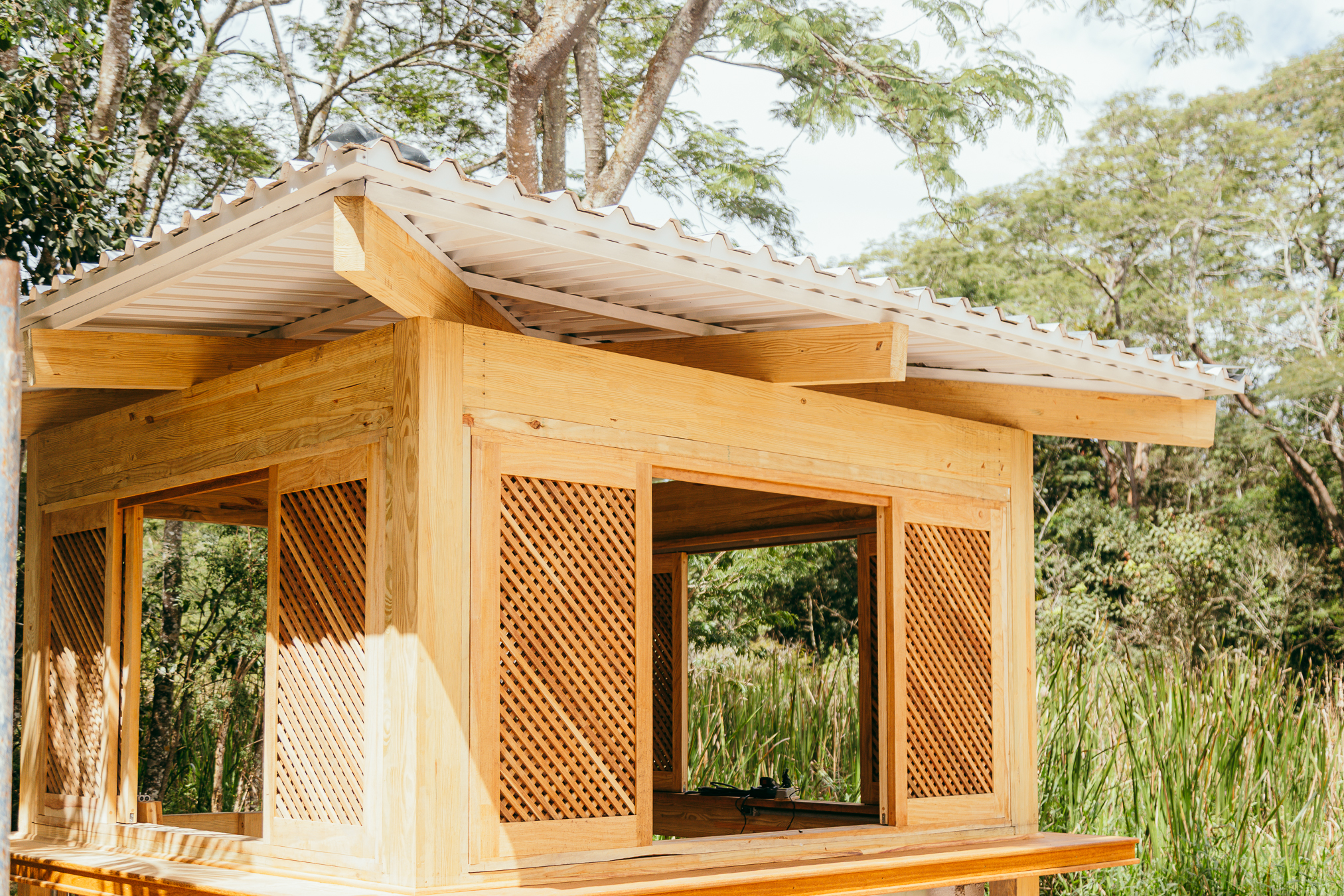 Pequeno quiosque de madeira clara, com paredes em treliça e telhado inclinado, cercado por árvores e vegetação verde, iluminado pelo sol.