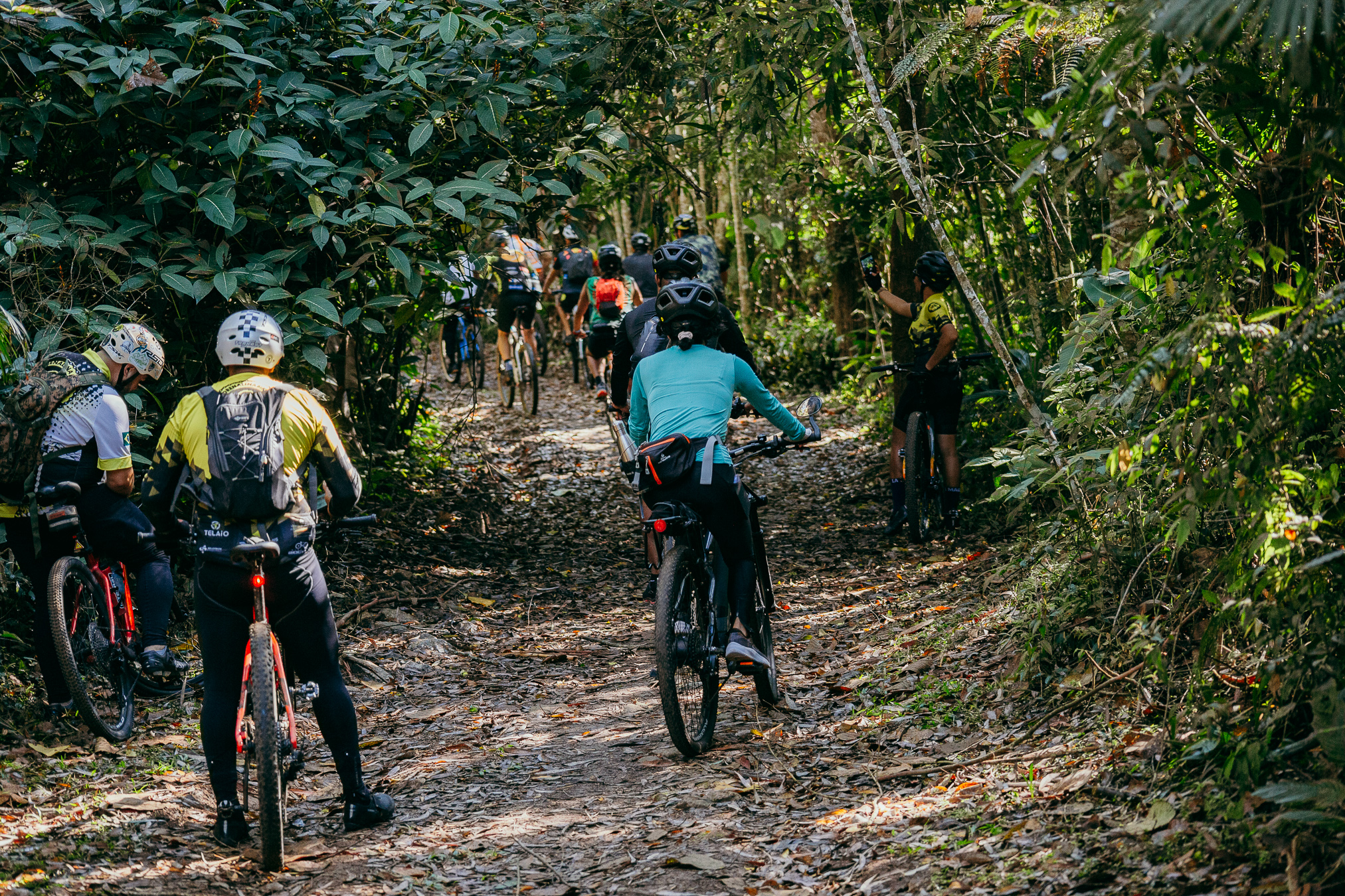 A imagem mostra um grupo de ciclistas percorrendo uma trilha estreita dentro de uma floresta bastante fechada.  No primeiro plano, há três ciclistas. À esquerda, dois estão próximos um do outro, usando capacetes e roupas esportivas predominantemente claras e escuras. Um deles carrega uma mochila. No centro, uma ciclista vista de costas veste uma camisa azul-clara, calça escura e capacete preto, pedalando uma bicicleta de montanha.  Mais à frente, há vários outros ciclistas seguindo pela trilha, alguns parcialmente escondidos pela vegetação. À direita, uma pessoa está parada ao lado da trilha, também com bicicleta, observando o grupo.  A trilha é de terra, coberta por folhas secas e pequenos galhos. A vegetação é muito densa, com árvores, arbustos e folhas verdes formando uma espécie de túnel natural sobre o caminho. A luz do sol atravessa a copa das árvores e ilumina partes da trilha, criando áreas alternadas de sombra e claridade.  A cena transmite a sensação de um passeio coletivo de mountain bike em meio à natureza.