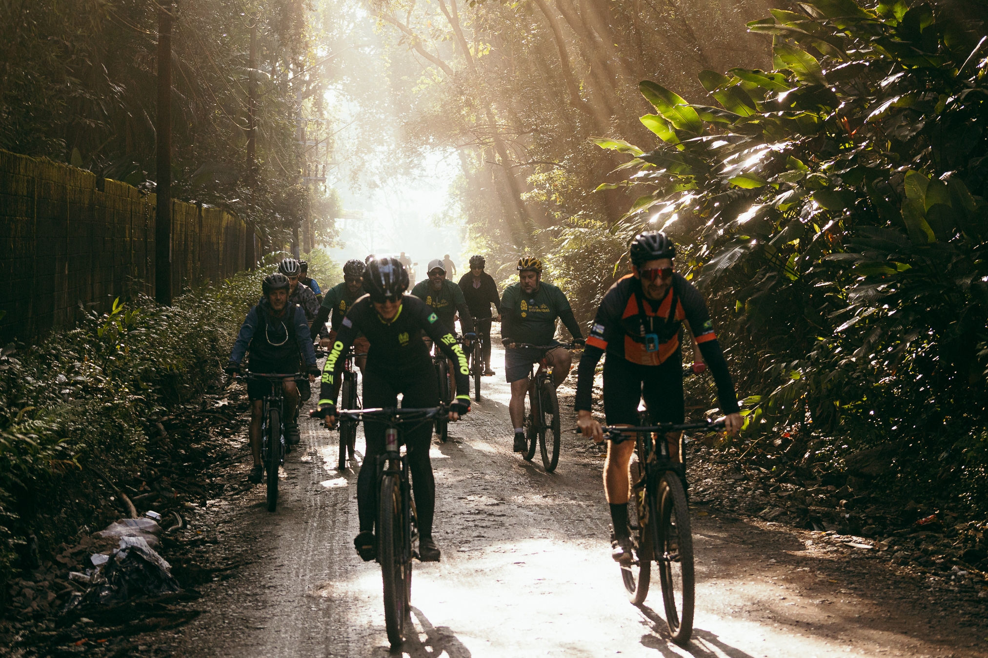 A imagem mostra um grupo de cerca de dez ciclistas pedalando por uma estrada estreita de terra, dentro de uma floresta densa.  Em primeiro plano, há dois ciclistas: um ao centro, vestido de preto e com capacete, e outro à direita, usando roupa esportiva preta e laranja. Atrás deles, vários outros ciclistas seguem pelo mesmo caminho.  A floresta é muito verde e fechada, com árvores altas dos dois lados e bastante vegetação rasteira. À esquerda, há uma cerca feita de bambu ou madeira. A luz do sol atravessa as copas das árvores, formando feixes luminosos e deixando o fundo da estrada muito claro. O ambiente parece úmido e tranquilo, apesar do grupo estar em movimento.  A fotografia transmite a sensação de um passeio de bicicleta em meio à natureza, com forte contraste entre as áreas sombreadas da floresta e a luz dourada do sol.