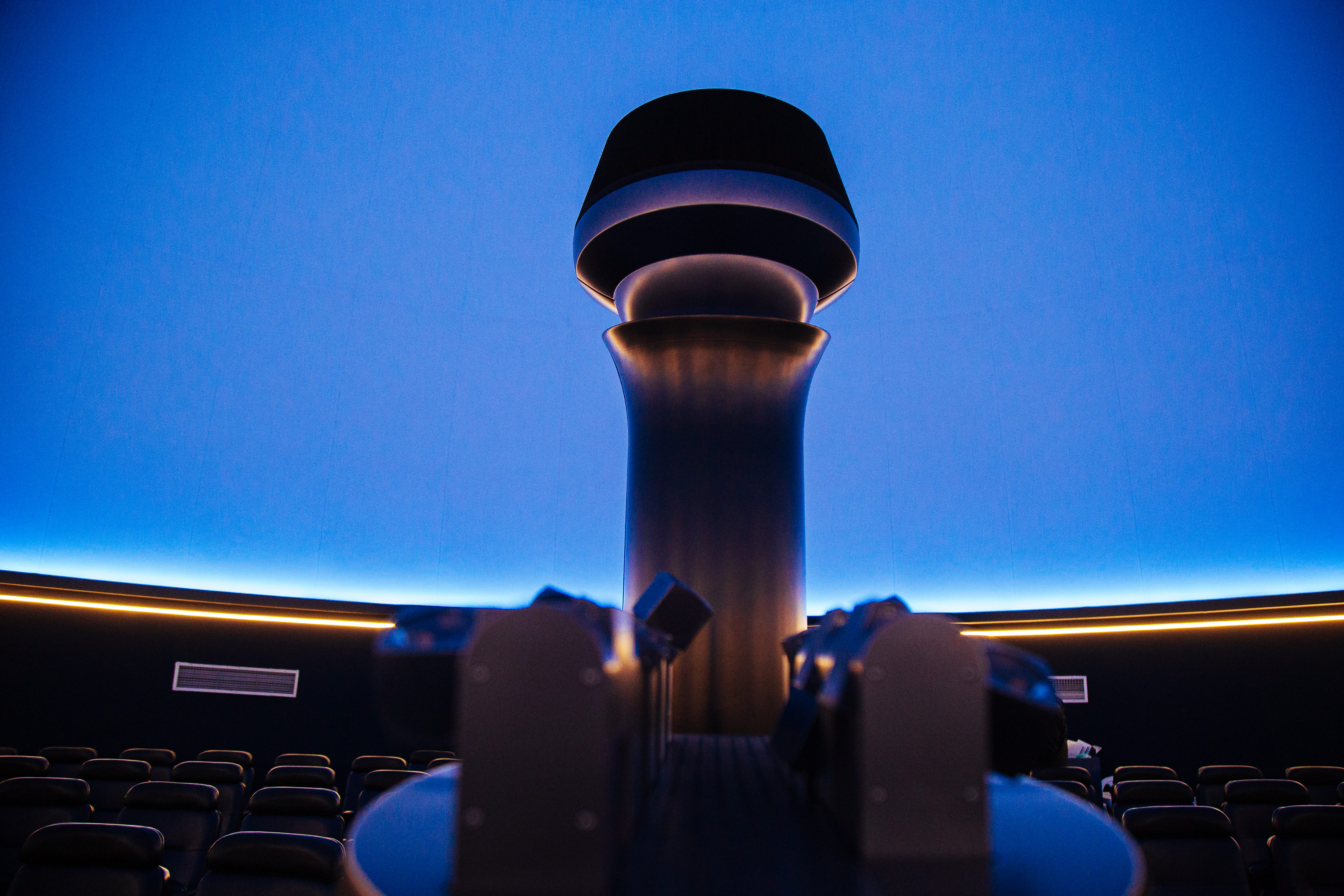Imagem mostra o interior de um planetário com cúpula azul, projetor central e poltronas reclináveis em semicírculo. Espaço acessível e imersivo, voltado à educação astronômica.