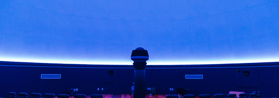 A imagem mostra o interior de um planetário. O teto arredondado iluminado por uma luz azul cria uma atmosfera imersiva para projeções astronômicas. No centro, há um projetor esférico característico, usado para exibir imagens do espaço e das estrelas. O ambiente é composto por cadeiras dispostas ao redor, proporcionando uma experiência confortável para os espectadores. O design escuro das paredes e do chão ajuda a minimizar reflexos e maximizar a visualização das projeções.